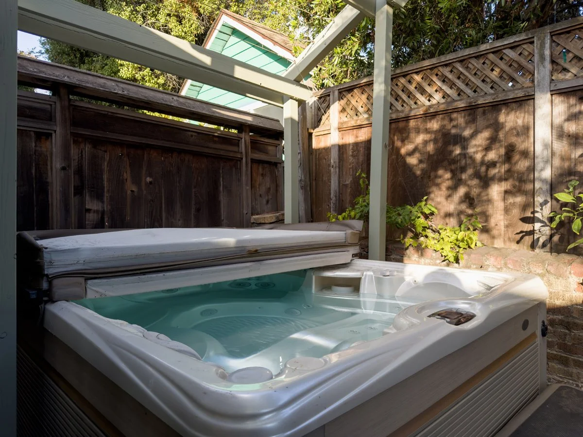 An outdoor hot tub with a cover partially open, surrounded by a wooden fence and garden plants, with a small house or shed partially visible in the background under a canopy of trees.
