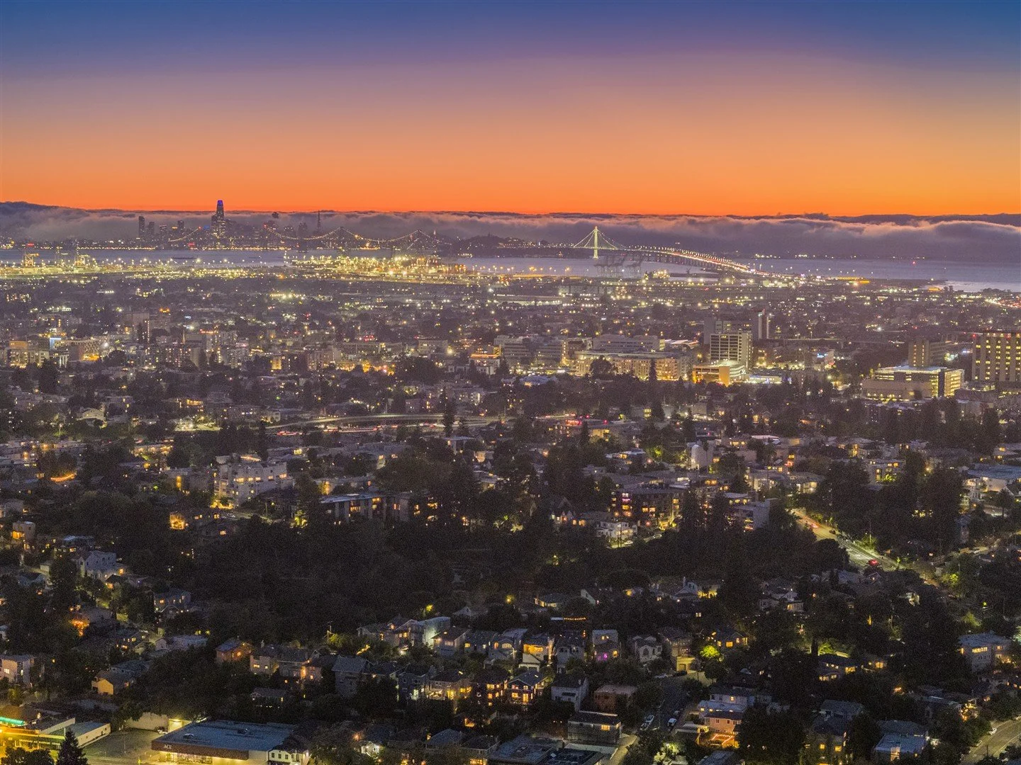 Cityscape at sunset with illuminated buildings, a bridge, and a view of the water against a colorful sky.