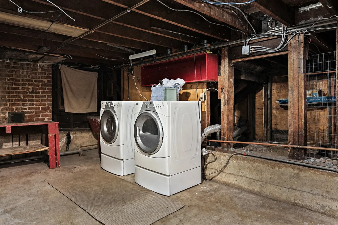 A basement laundry area with a brick wall, wooden ceiling beams, and two front-loading washing machines. There is a small red workbench on the left and various utilities and wiring visible on the ceiling and walls.