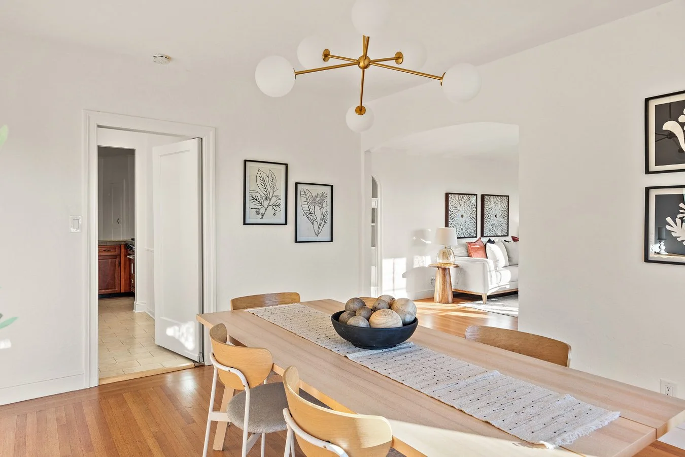 Dining area with a wooden table, a black bowl with round stones, and a decorative cloth runner. Adjacent living room with white sofa and framed artwork, in a modern, minimalist style.