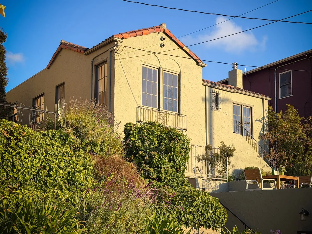 A two-story yellow house with a red tiled roof, large bay windows, and surrounded by lush green plants and shrubs, with a small patio area with chairs and a table in front.