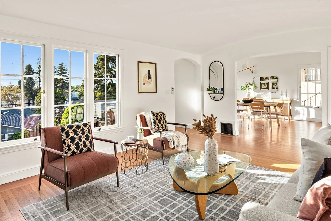 Bright living room with large windows, two pink armchairs with patterned pillows, a glass coffee table with vases and books, and a beige sofa with pillows, leading to a dining area with a wooden table and chairs.
