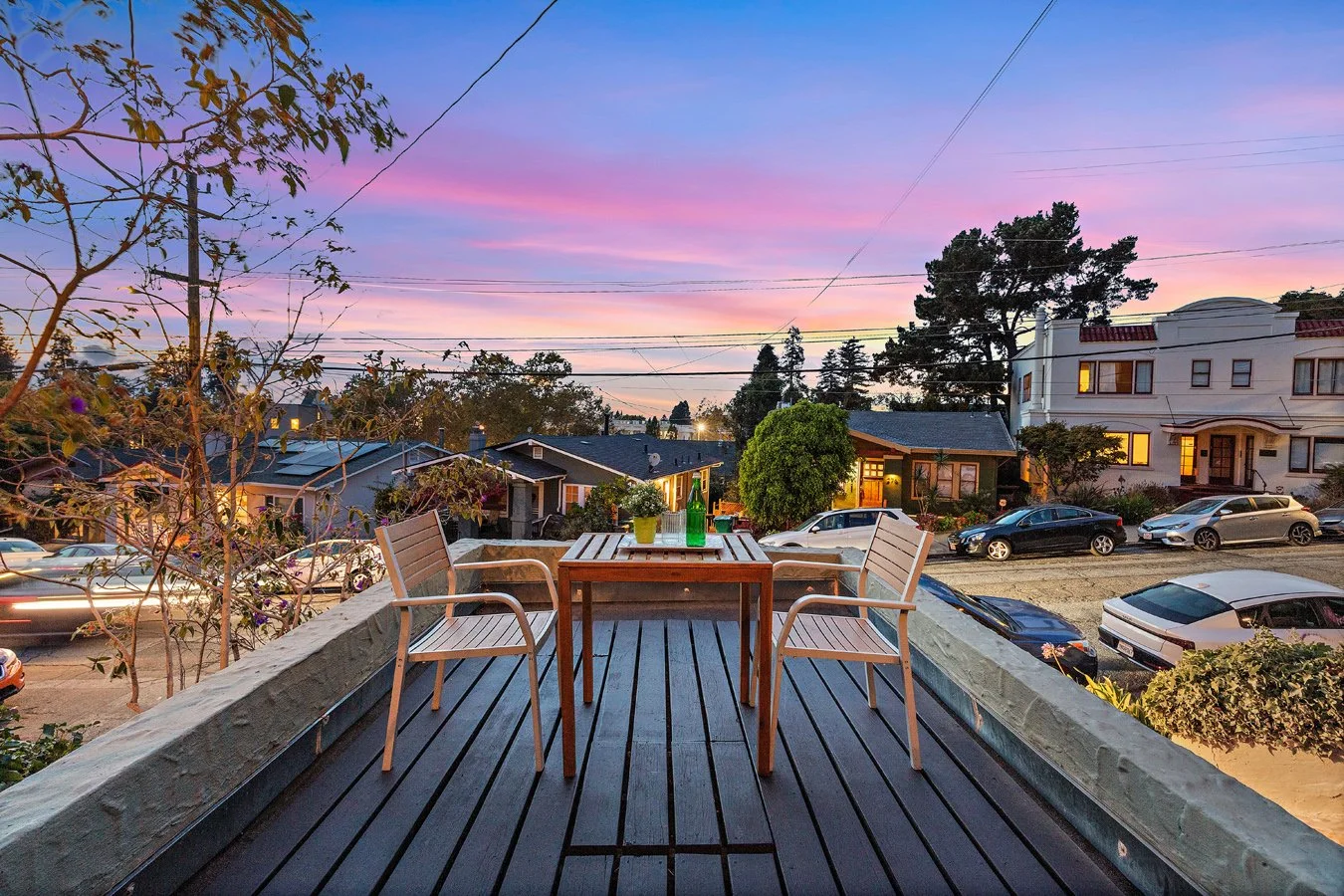 View from a balcony showing a wooden table with four chairs, potted plants, and a cityscape with houses, parked cars, and trees during sunset with pink and purple sky.
