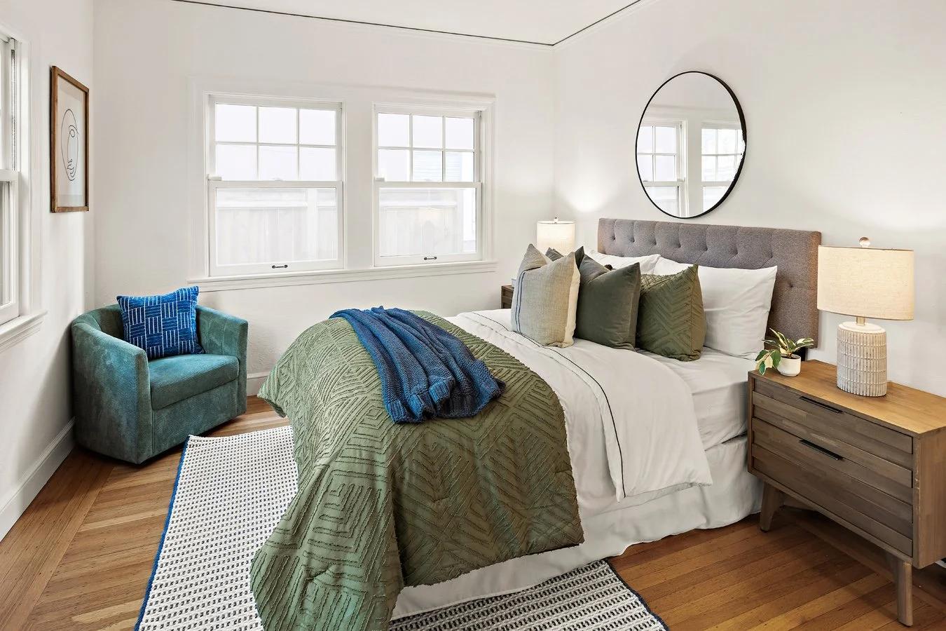 A cozy bedroom with a bed covered in white sheets, green and beige pillows, and a green quilt. Next to the bed is a wooden nightstand with a beige lamp and a small potted plant. To the left, a teal armchair with a blue patterned pillow is beside two 