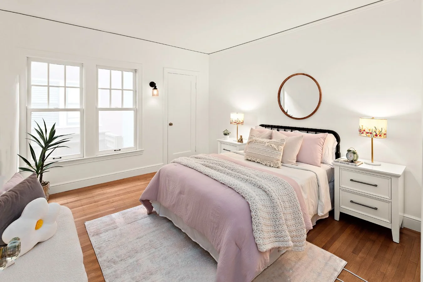 Bedroom with white walls, hardwood floor, a bed with light pink bedding, two white nightstands with lamps, round mirror on the wall, window with white trim, a potted plant, and a woven blanket on the bed.