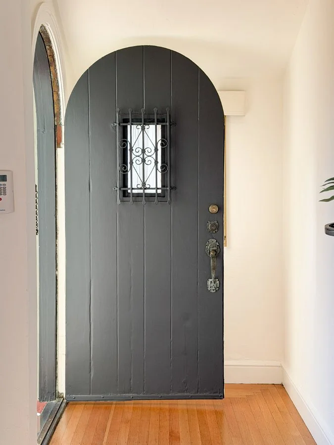 A black wooden front door with a small barred window near the top, a vintage-style handle, and a latch, inside a bright home with white walls and a wooden floor.