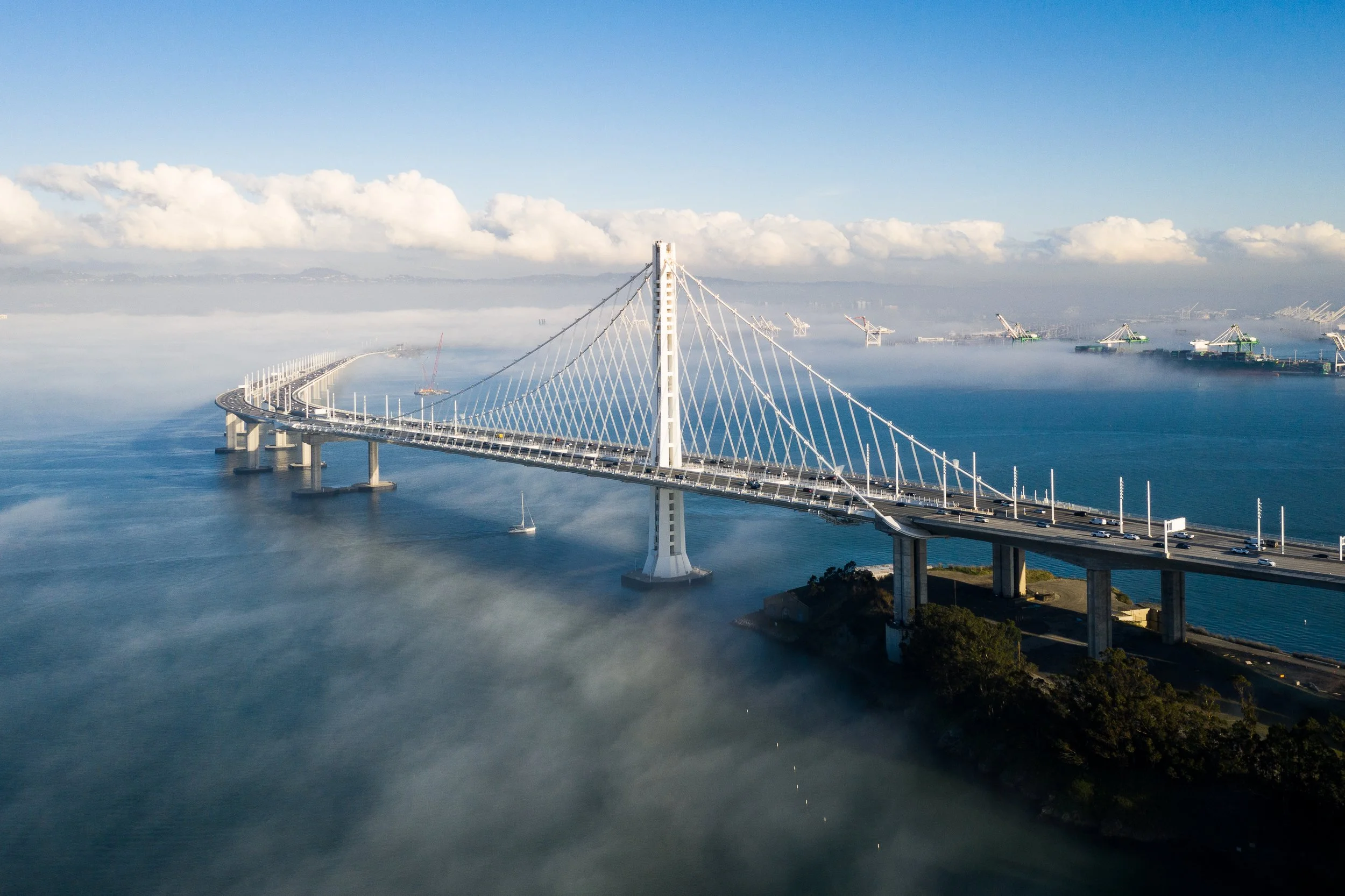 A large suspension bridge over a body of water with fog or mist beneath, cranes in the background, and a partly cloudy sky.