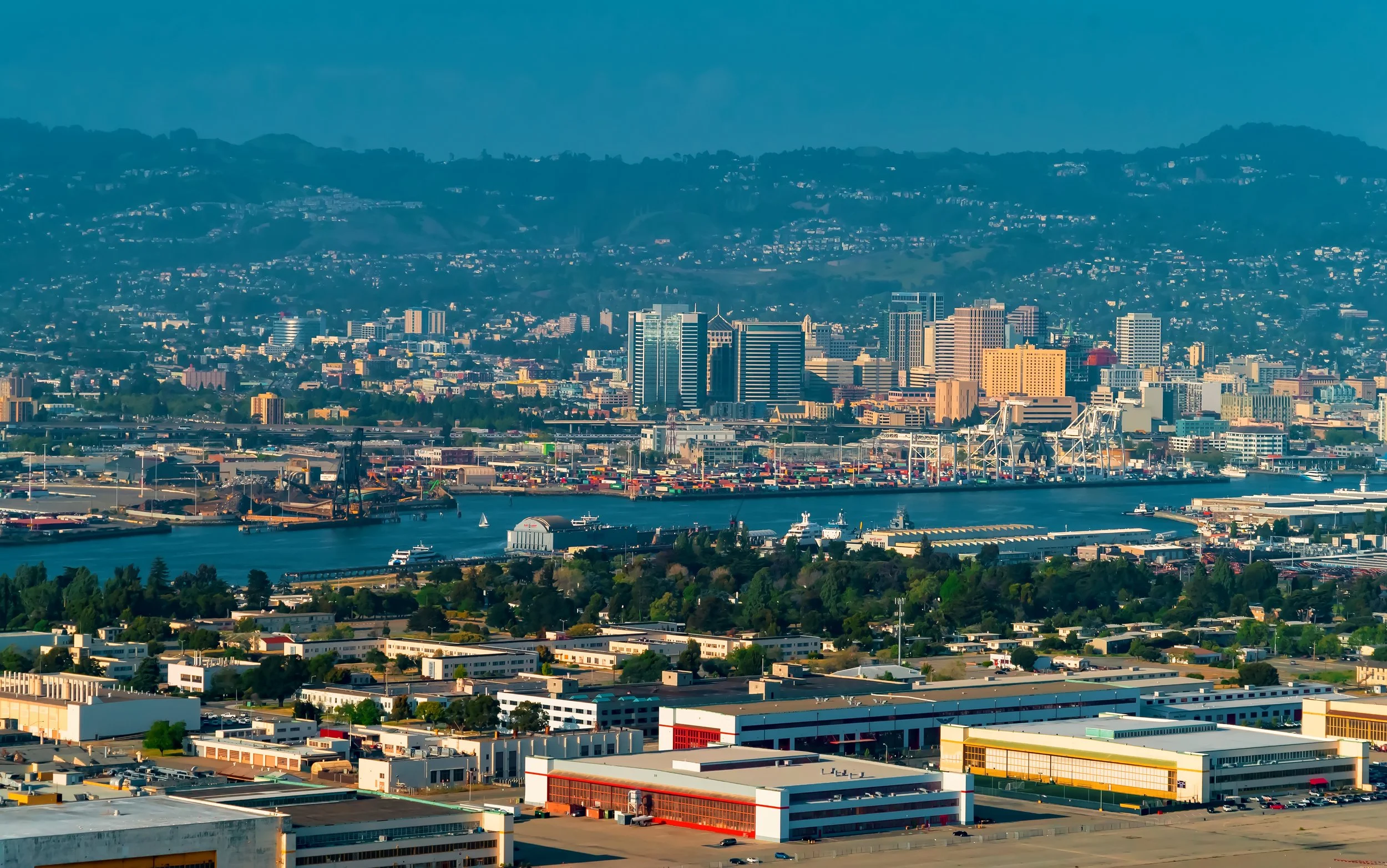 City skyline with high-rise buildings along a harbor with cargo containers and cranes, hills in the background under a clear sky.