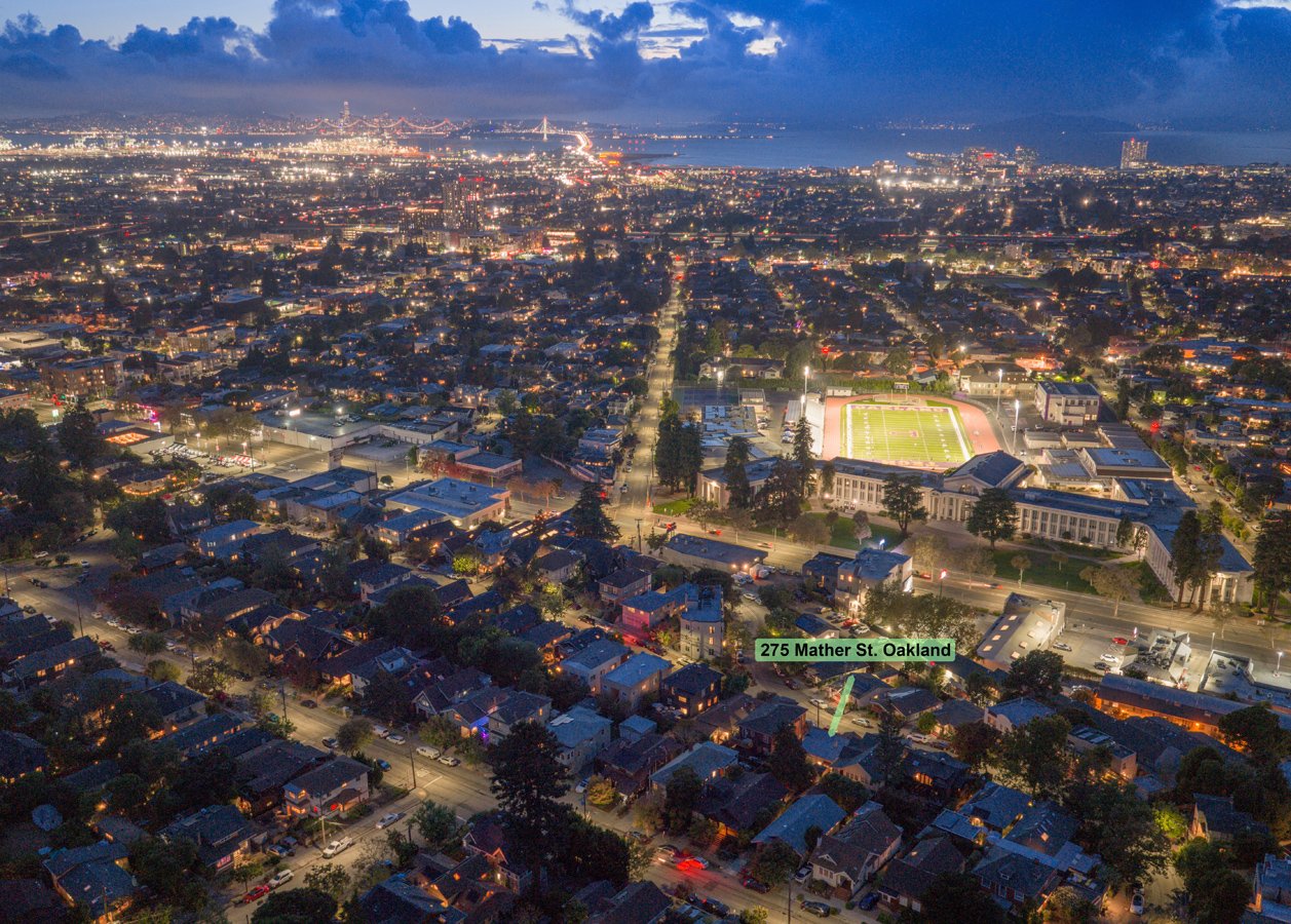Nighttime aerial view of Oakland, California, featuring a grid of residential streets, a sports stadium, and the San Francisco Bay in the background, with illuminated bridges and city lights.