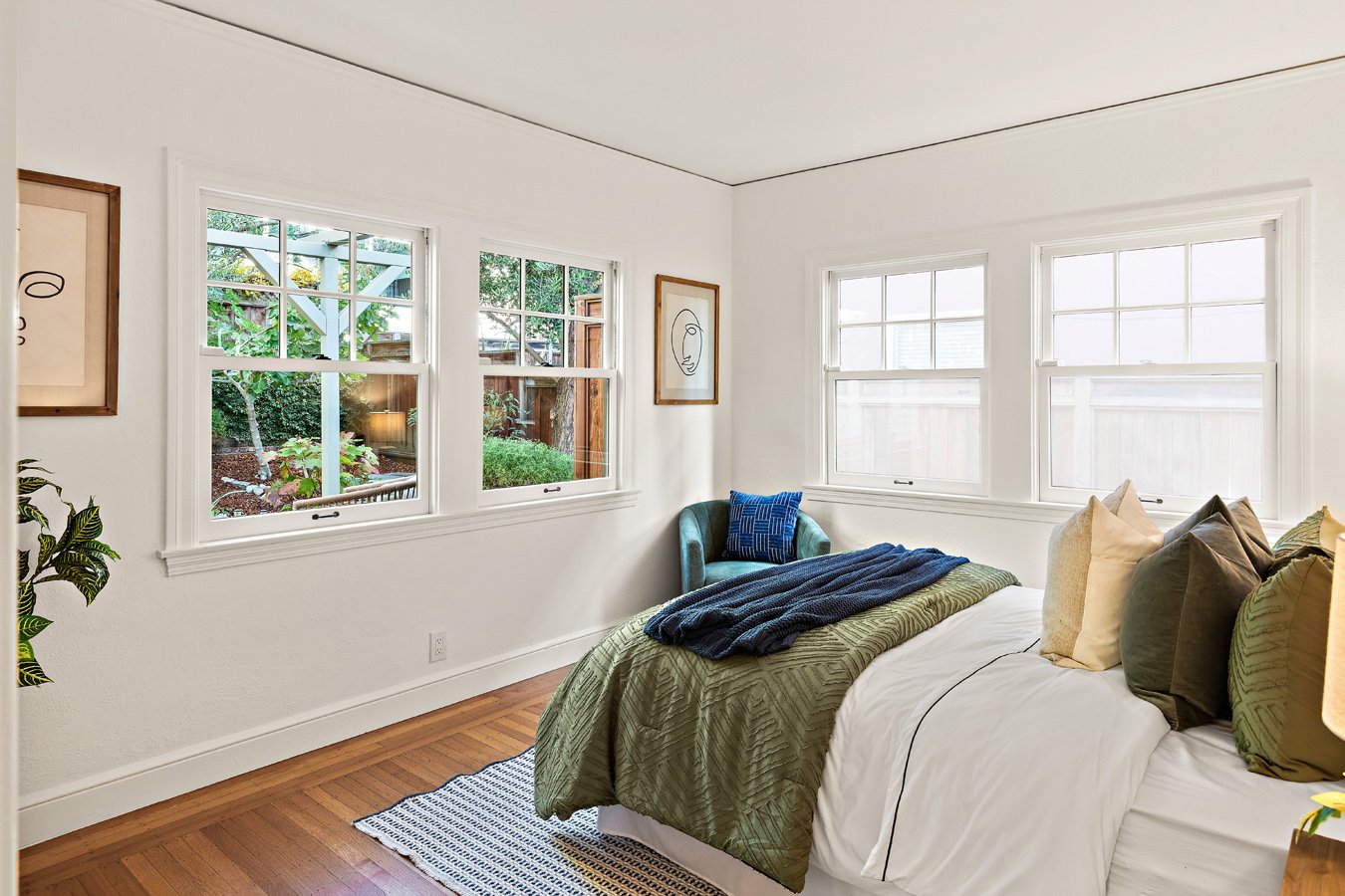 A bright bedroom with white walls, large windows, a bed with multiple pillows, a green and white comforter, a blue throw blanket, and a blue armchair near the corner.