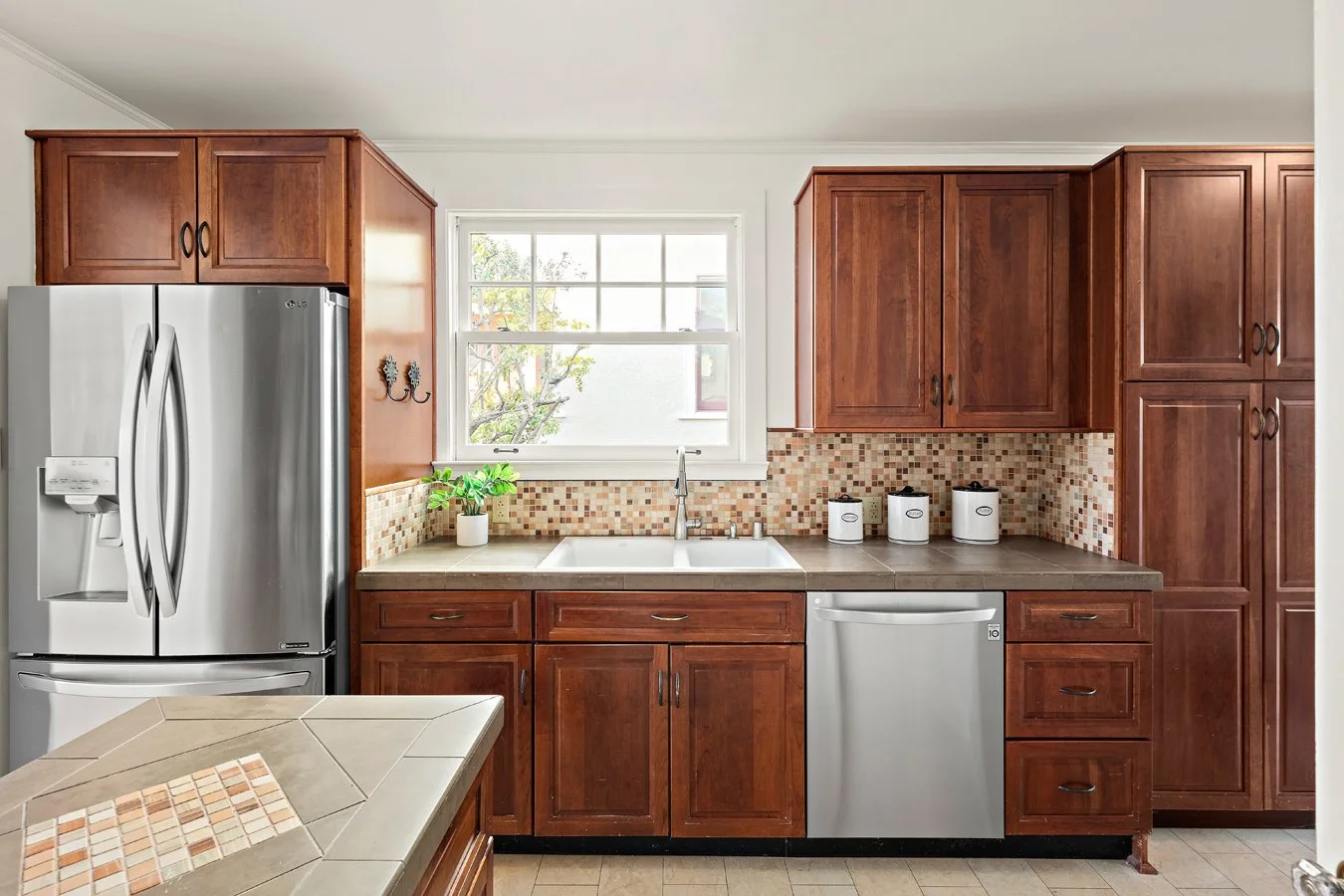Kitchen with wooden cabinets, stainless steel refrigerator, dishwasher, sink, window, and countertop with small containers and potted plant.