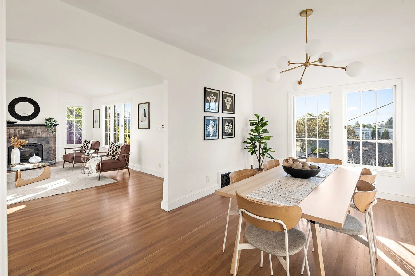 A bright, modern dining area with wooden chairs around a rectangular table, a black bowl with decorative objects, and large windows showing a suburban neighborhood. Adjacent living area with two chairs, a fireplace, and wall art.
