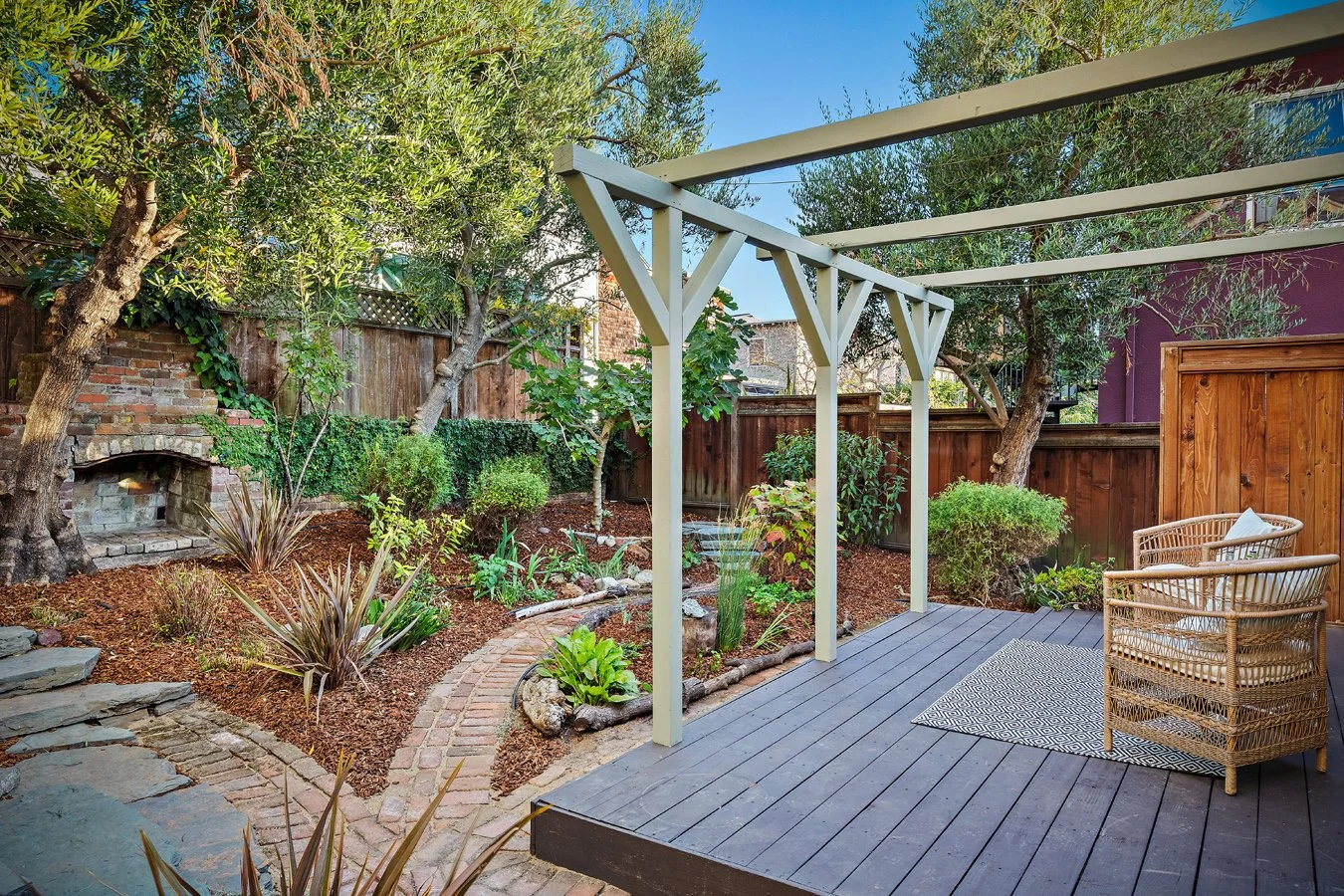 Backyard patio with a wooden deck, two wicker chairs with cushions, a rug, and a garden with trees, shrubs, and a brick pathway.