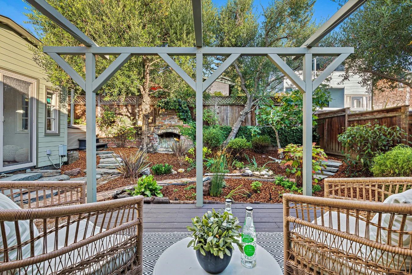 View of a backyard garden with trees, plants, and a stone pathway from a screened porch area with outdoor wicker furniture and a small table.