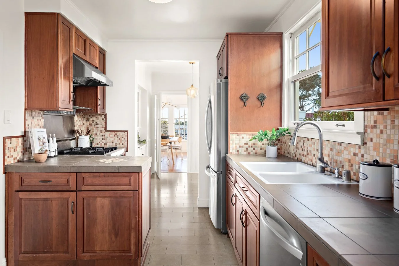 A bright kitchen with wooden cabinets, beige tiled countertops, a stainless steel refrigerator, a white double sink, and a window overlooking outdoors, with a view into a dining area with a chandelier and chairs.