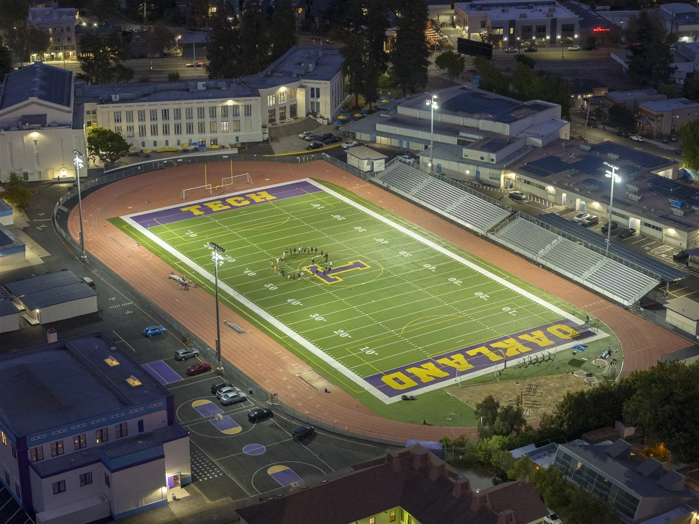 An aerial view of a football field at night, illuminated with bright lights. The field has the word 'TECH' painted in large yellow letters on a purple background at each end zone, and the perimeter features a running track. Surrounding the stadium ar