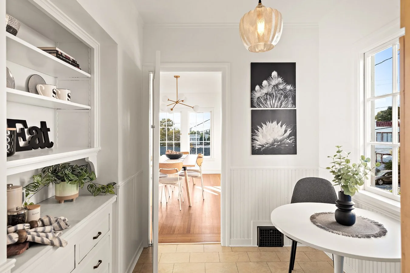 A bright dining area with white walls, a small white table with a black vase and green plant, gray chair, and large windows. There is wall art with black and white floral images, a round woven ceiling light, and a doorway leading to another room with