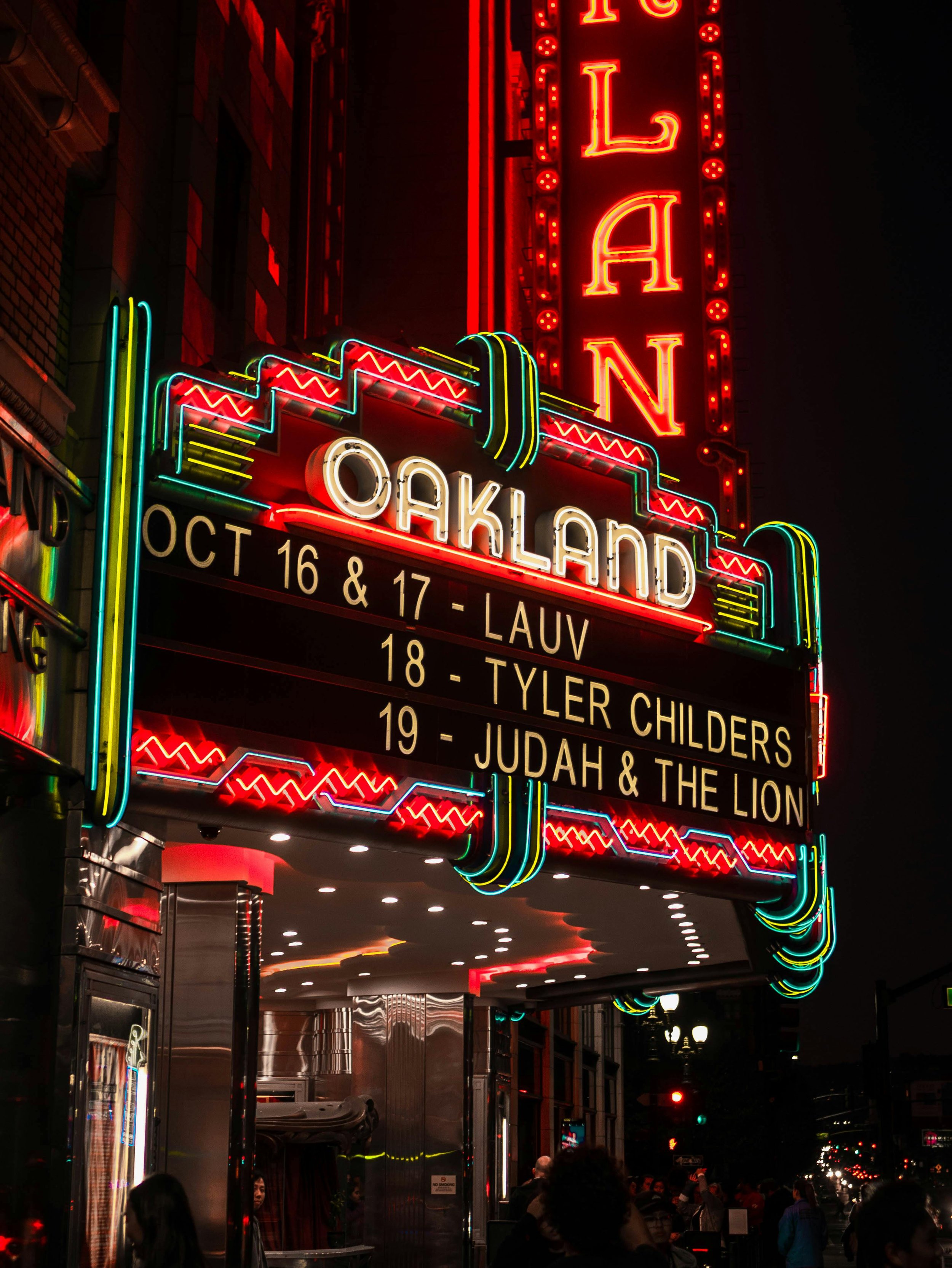 Neon sign for the TallOakland theater at night, with a digital marquee displaying a performance schedule and a crowd of people in front.