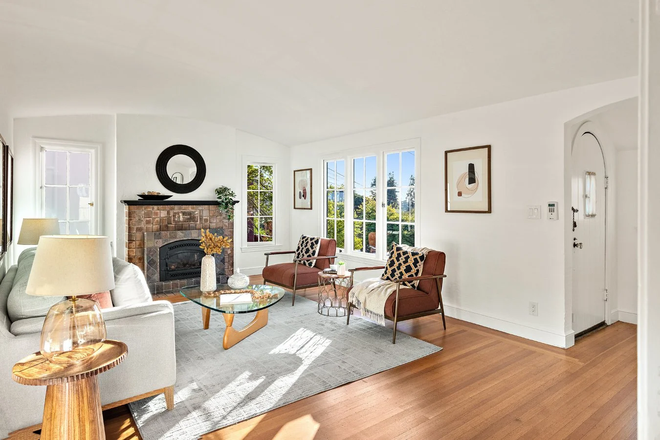 Bright living room with white walls, large windows, brick fireplace, beige sofa, two maroon armchairs, glass coffee table, and modern abstract artwork.