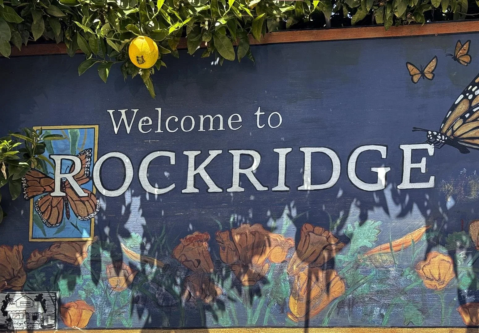 A colorful sign reading 'Welcome to Rockridge' surrounded by painted flowers and butterflies, with greenery and a yellow fruit at the top