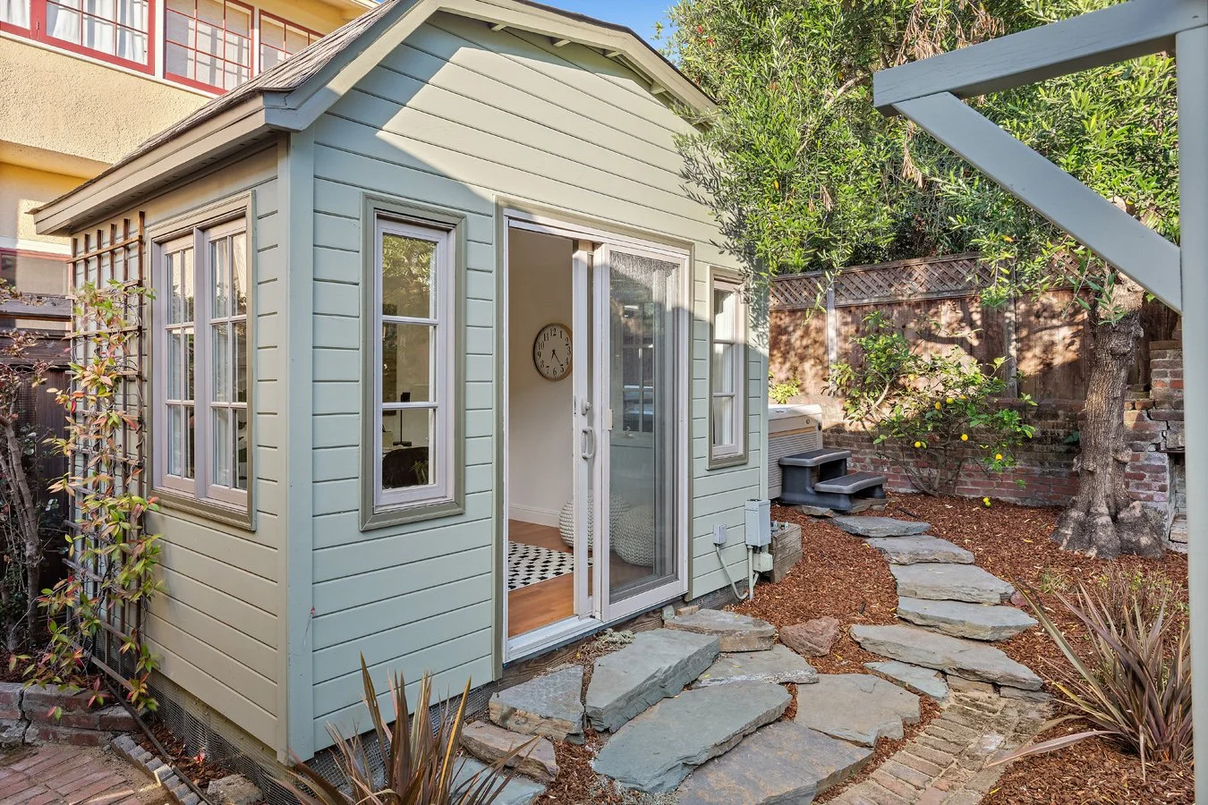Small light green shed with white trim and glass sliding door, situated in a backyard with stone pathway and trees.