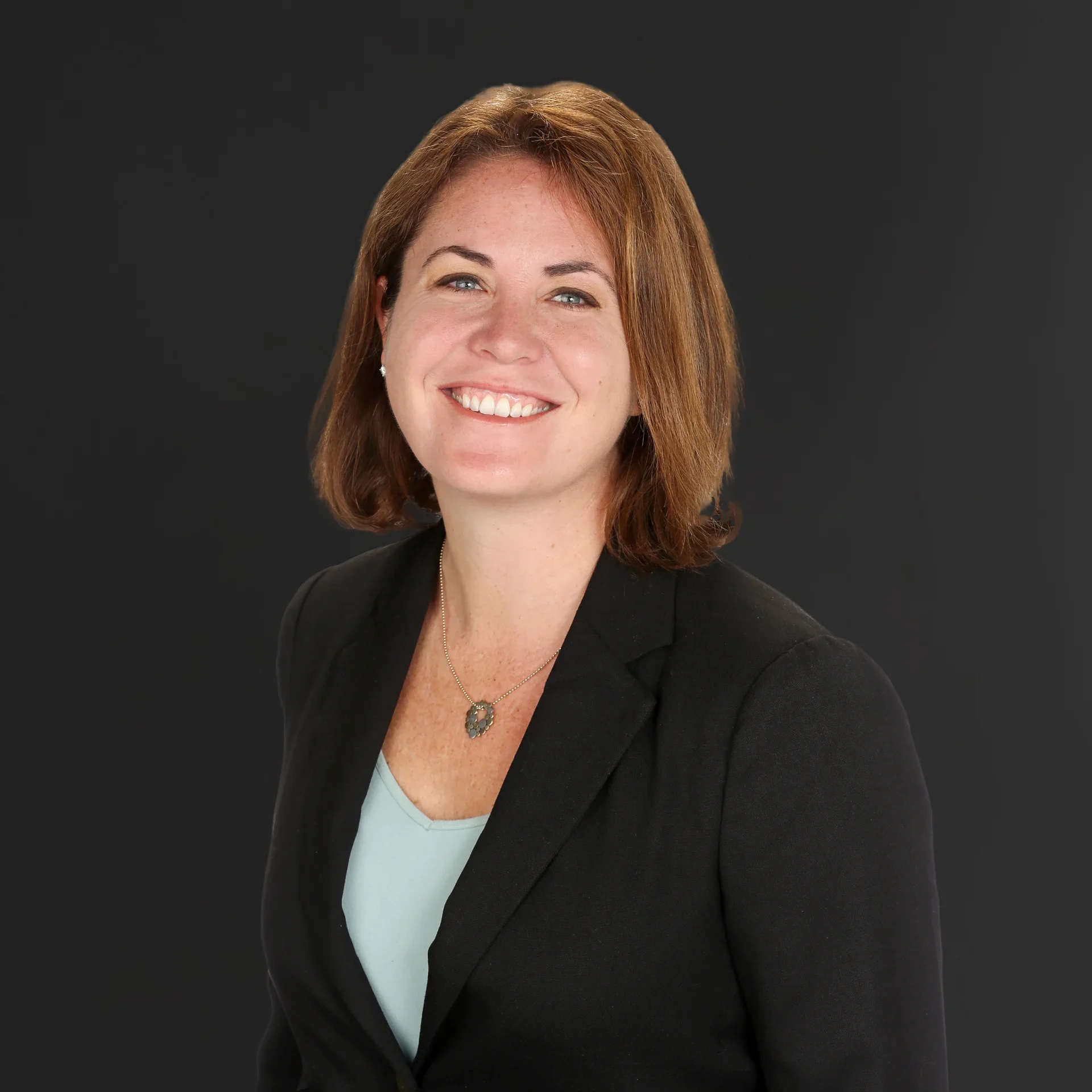 Headshot of a smiling woman with shoulder-length red hair, wearing a black blazer over a light blue top, against a dark background.