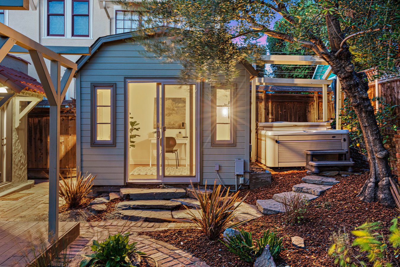 A backyard at dusk with a small, modern gray shed with glass sliding doors and windows, surrounded by a landscaped garden with stepping stones, plants, and a large tree, and a hot tub on the right side.