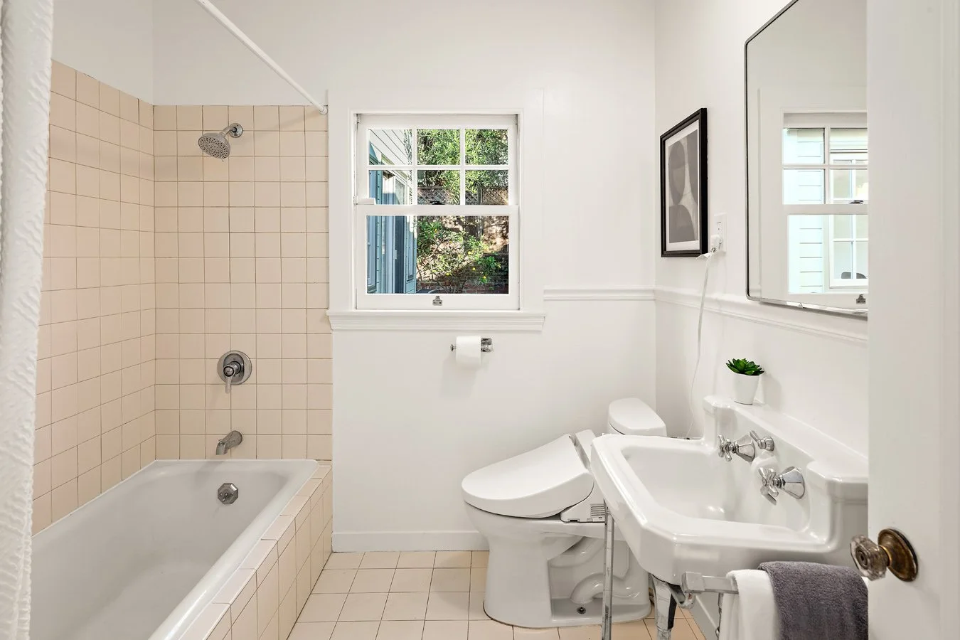 A small bathroom with a bathtub, shower, toilet, and sink, with white walls, beige tiles, and a window showing greenery outside.