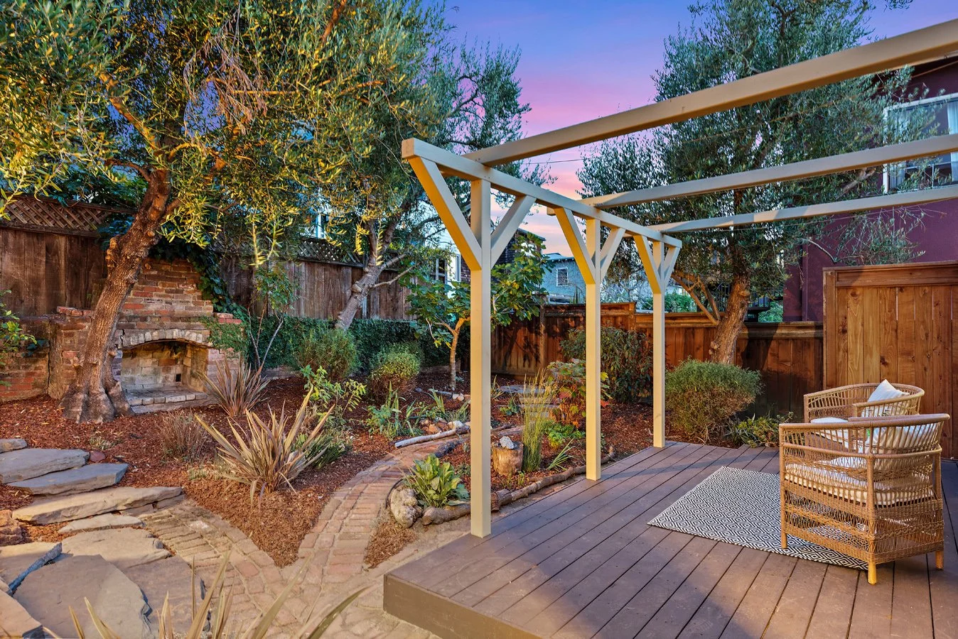 Backyard patio with wooden deck, wicker chairs, and a small rug. Surrounding garden with trees, shrubs, and a brick pathway. A wooden fence and a brick outdoor fireplace are visible in the background. The sky is colorful, indicating dusk or dawn.
