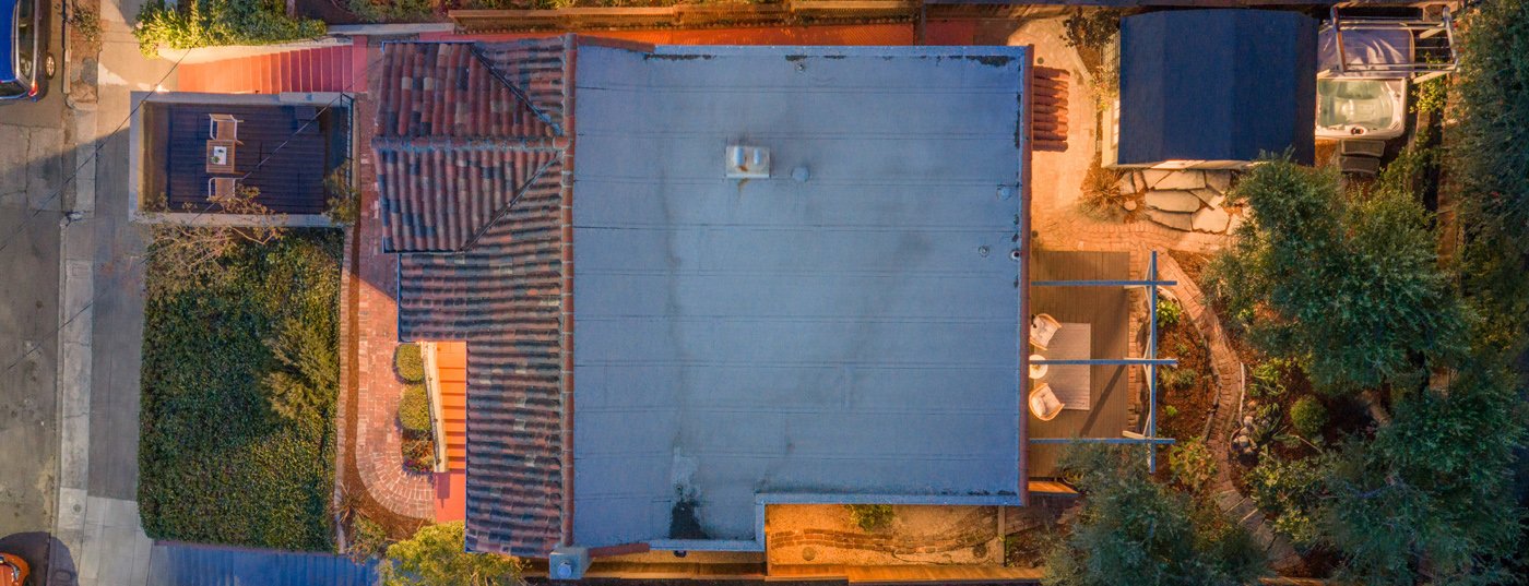 Aerial view of a house with a grey roof, a red brick chimney, surrounding trees, a small yard with an outdoor table and chairs, and neighboring roofs and streets.