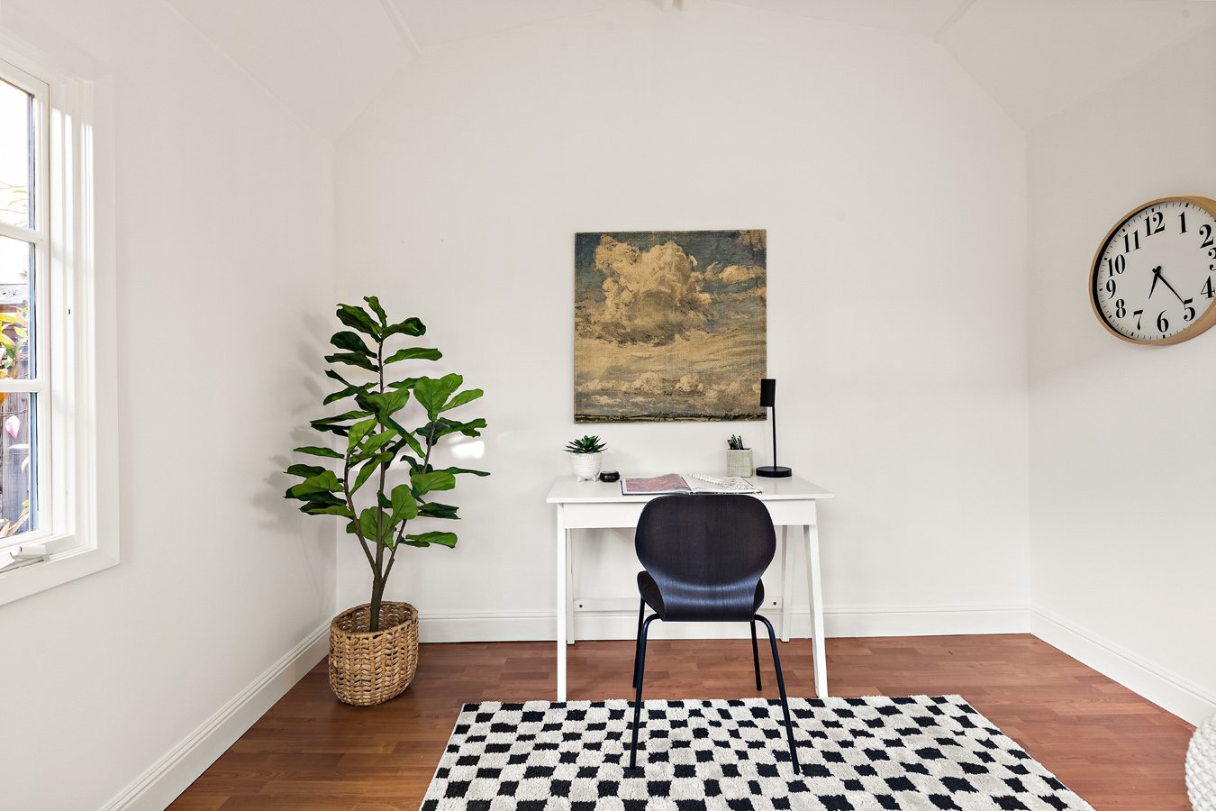 A minimalistic home office with a white desk, black chair, and decorative items, positioned against a white wall with a painting, clock, and a green potted plant nearby, beside a window with white frames, on a black and white patterned rug.