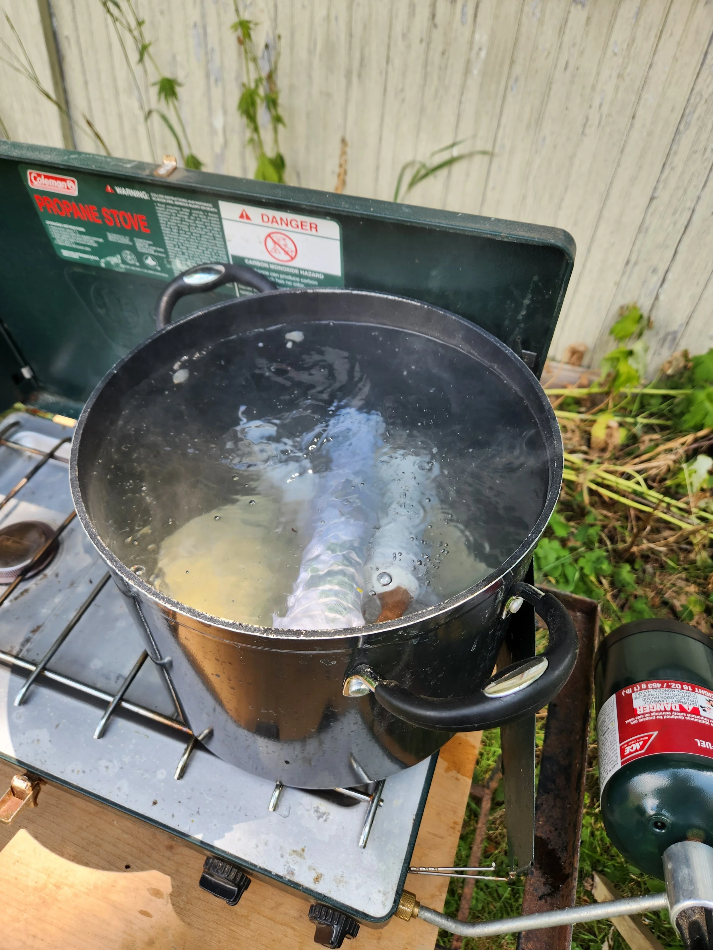 A black propane stove with water boiling inside, placed outdoors on a metal surface with a wooden board below. An empty propane tank is attached to the stove, and there is a wooden fence and some plants in the background.