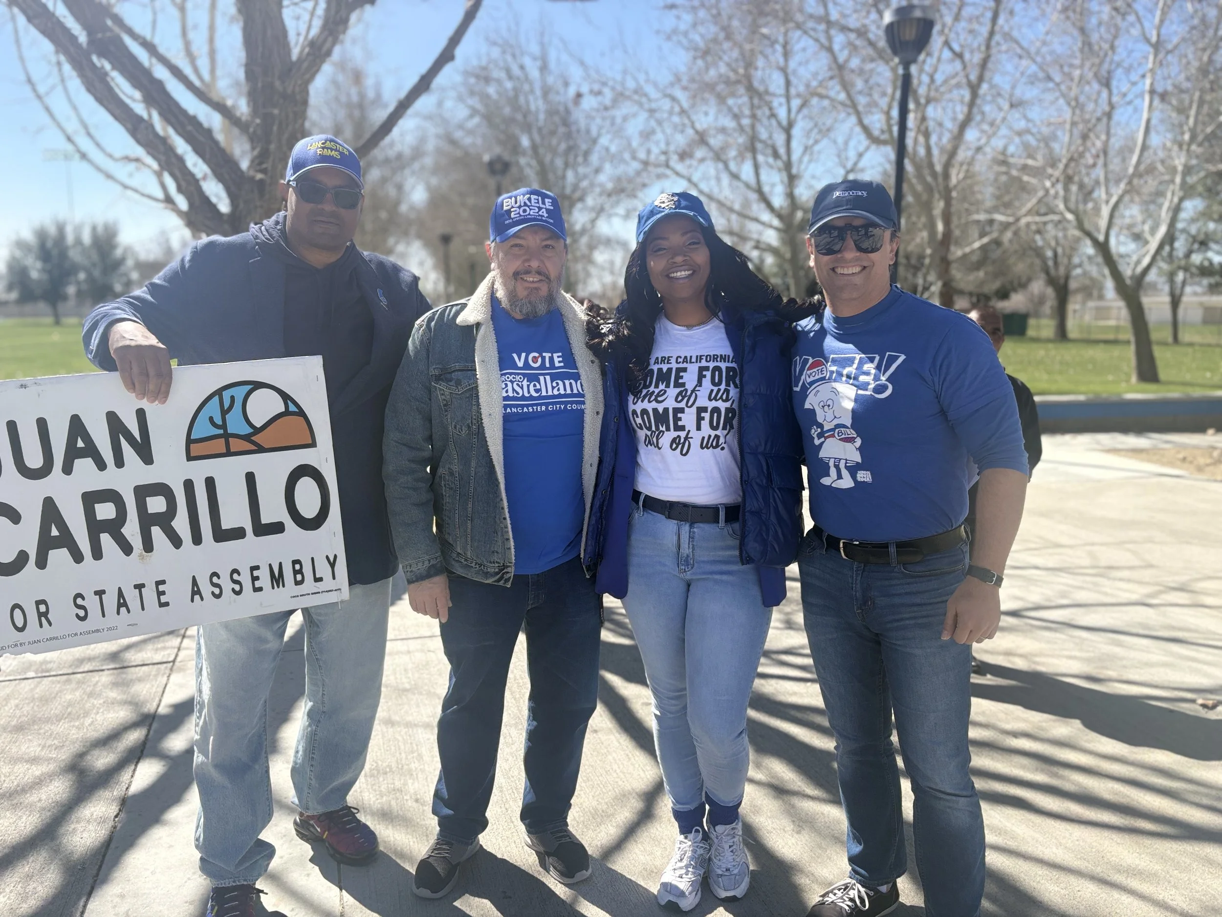 Four people standing outdoors during a sunny day, smiling and holding a large sign that says 'Juan Carrillo for State Assembly.' They are dressed casually, some wearing hats with campaign slogans. There are leafless trees and a grassy area in the background.