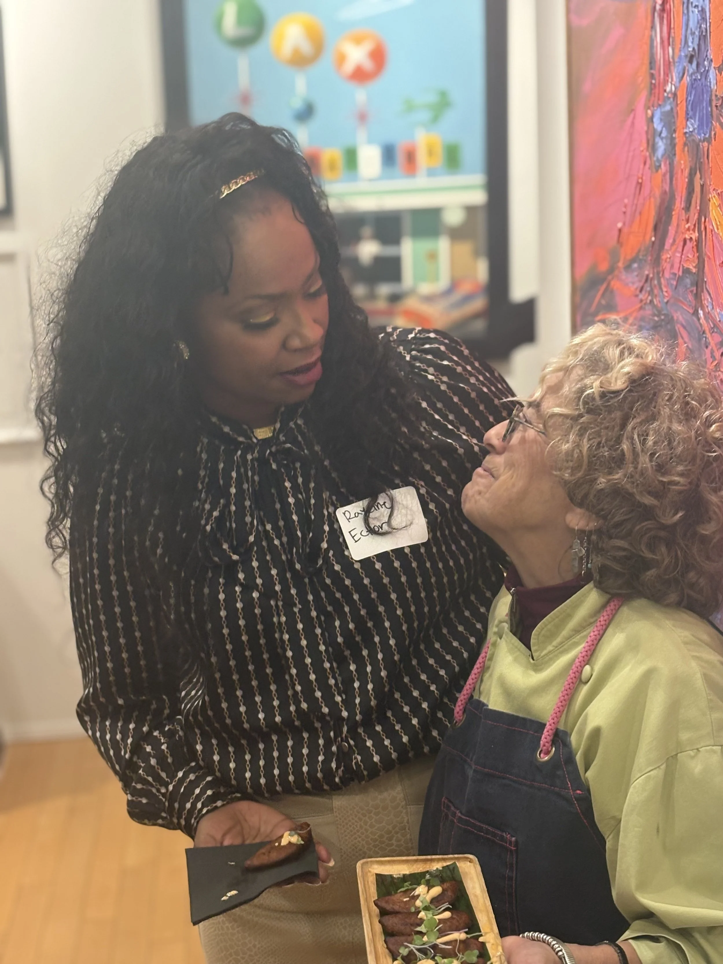 Two women are at an art gallery, one is giving the other a piece of chocolate. A tray of chocolates decorated with herbs is on a table between them.