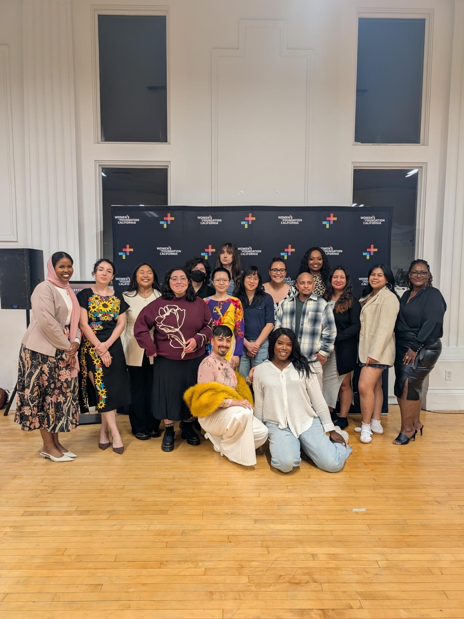 A diverse group of women and one man posing together in front of a backdrop with the Women's Foundation California logo. The group is standing in a room with wooden floors and large windows.
