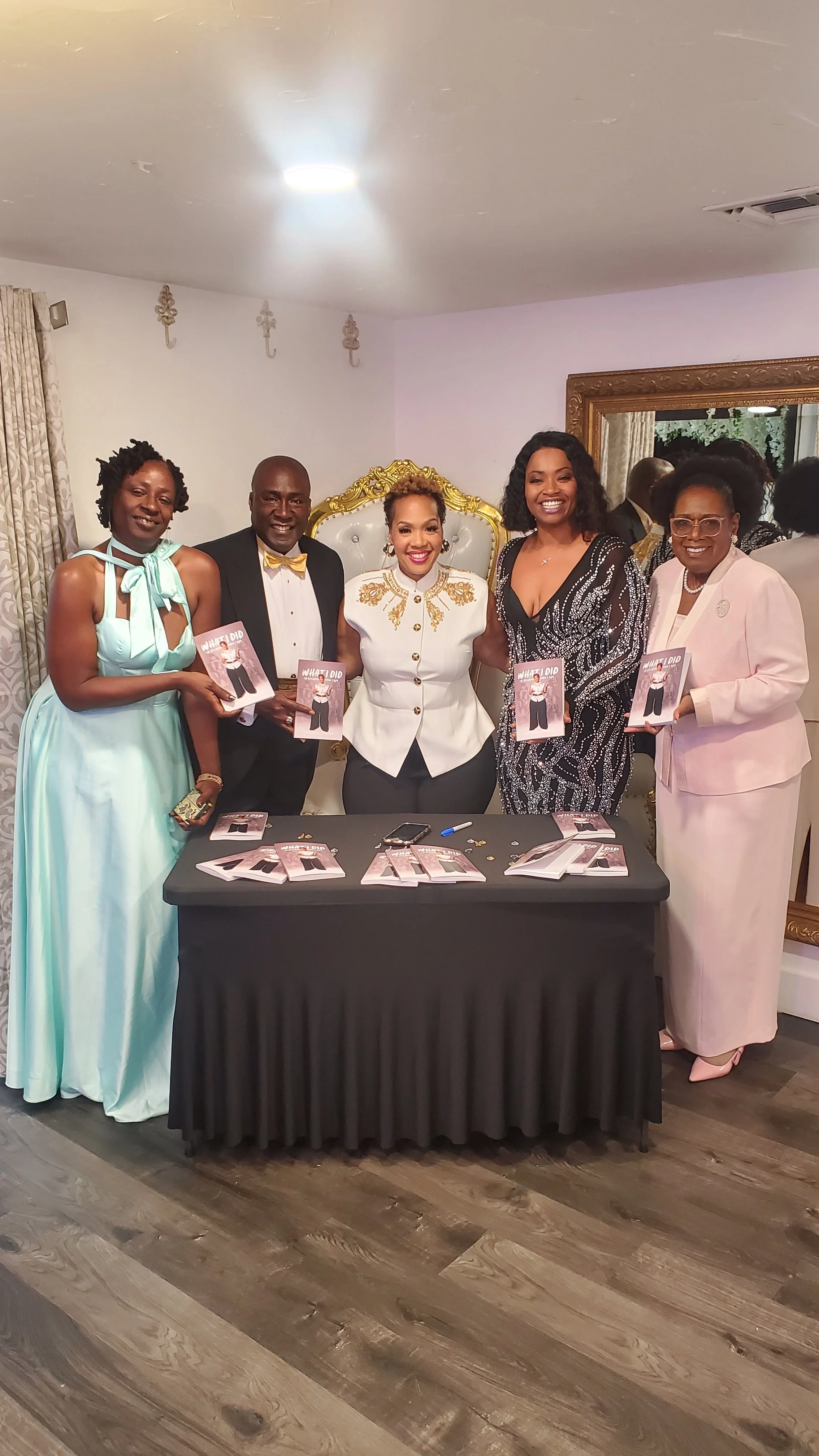 Group of five women and one man standing behind a table at a book signing event, holding copies of a book titled 'WHAT DID I DO'. The women are dressed in formal attire, including gowns and dresses, and the man is wearing a tuxedo with a bow tie. The setting appears to be a decorated indoor space with a mirror and elegant curtains.