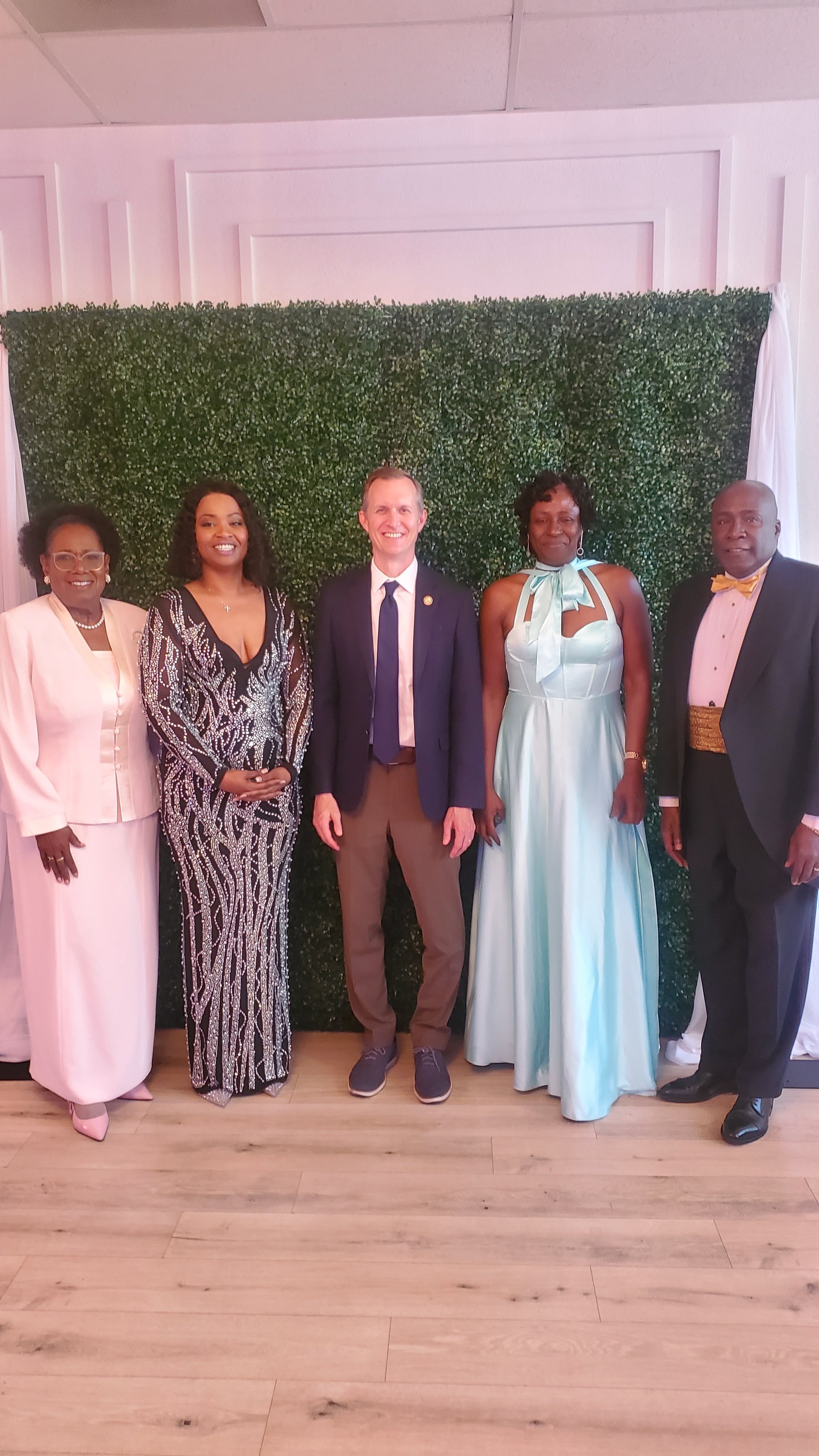 A group of six formally dressed people standing in front of a green hedge wall at an indoor event.