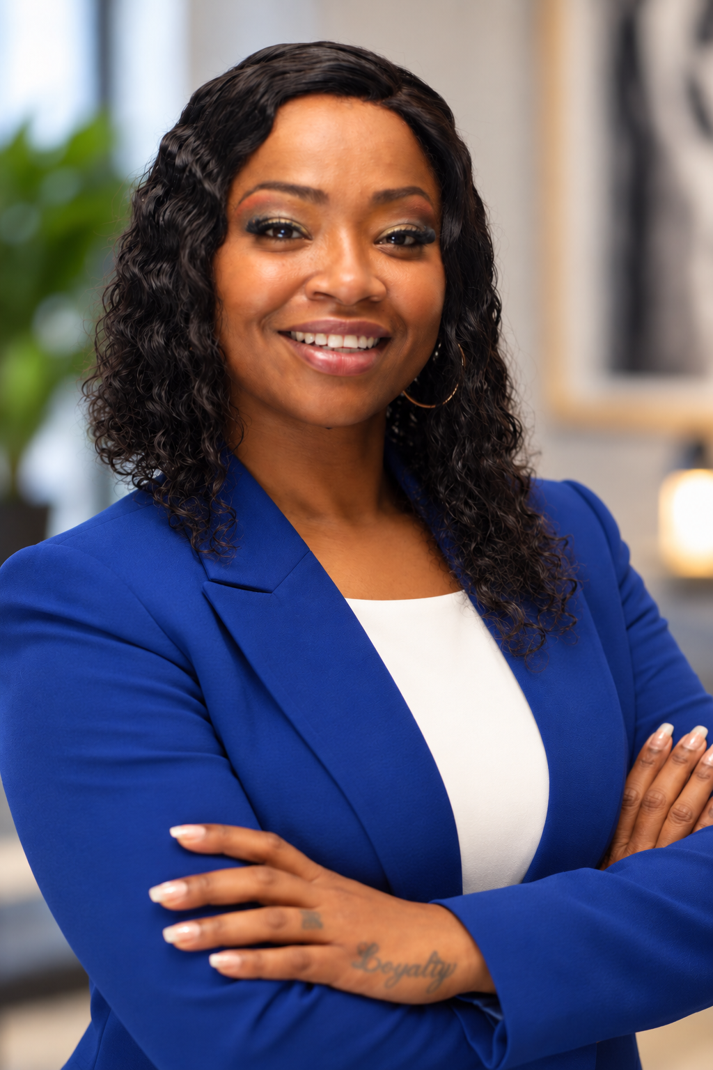 A smiling African American woman with curly hair, wearing a blue blazer and white top, standing with arms crossed in an office setting.