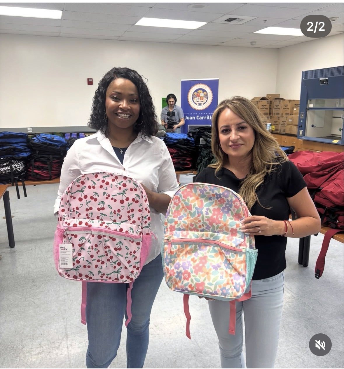 Two women standing in a room with stacks of backpacks behind them, holding colorful backpacks with floral patterns.