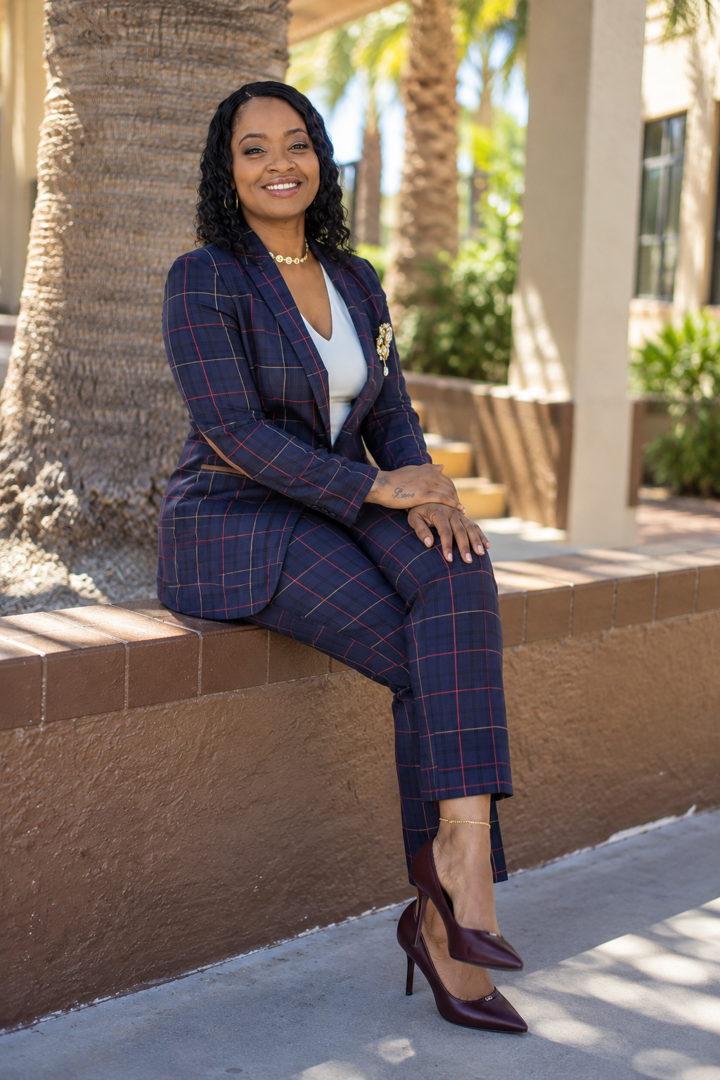 A woman sitting on a brick wall next to a tree outdoors, wearing a navy blue plaid suit, burgundy high heels, and jewelry, smiling at the camera.