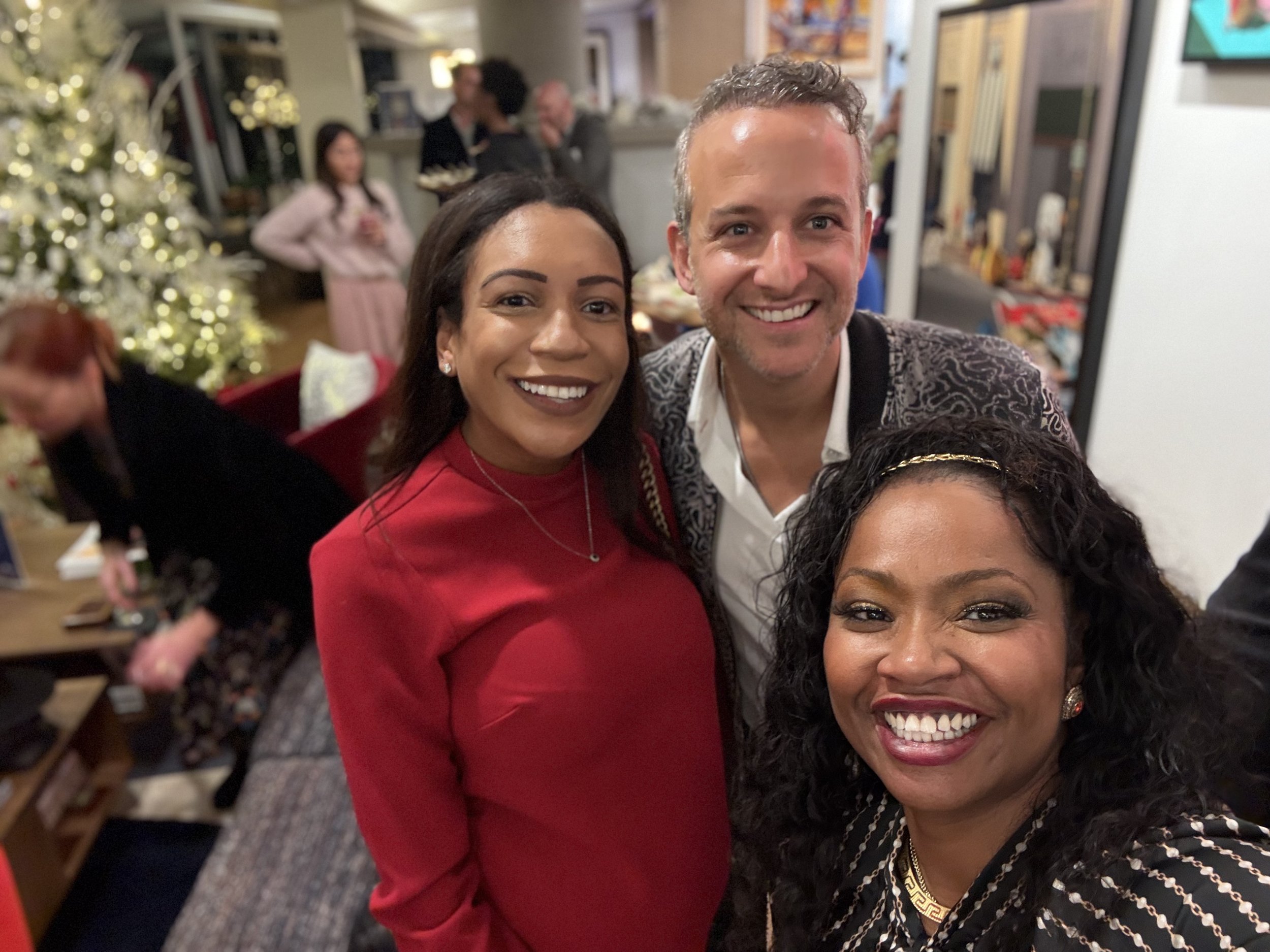 Smiling woman in red dress and smiling man in patterned sweater taking a selfie at a holiday gathering with Christmas tree and other guests in the background.