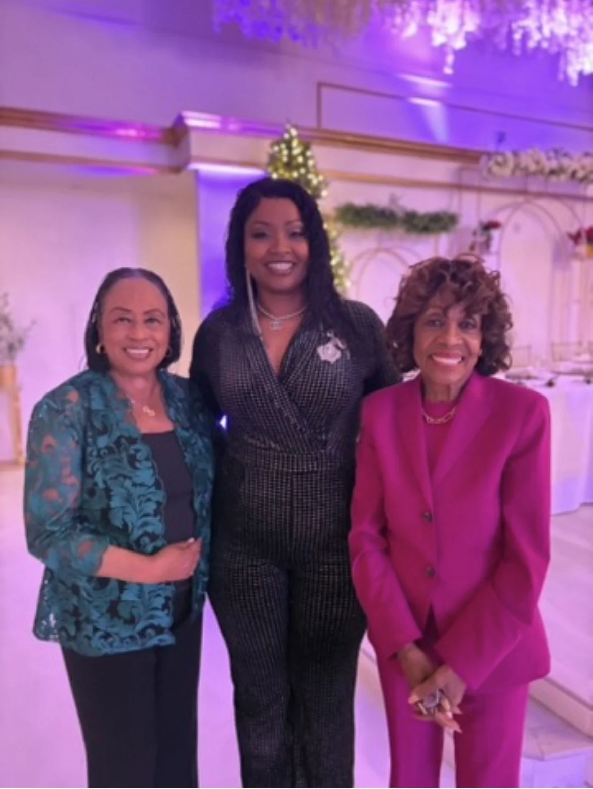 Three women standing indoors at a decorated event, smiling at the camera. The woman in the middle wears a black patterned outfit, the woman on the left wears a teal floral blazer, and the woman on the right wears a bright pink blazer with matching pants. The background features purple and white decorations, including a Christmas tree.