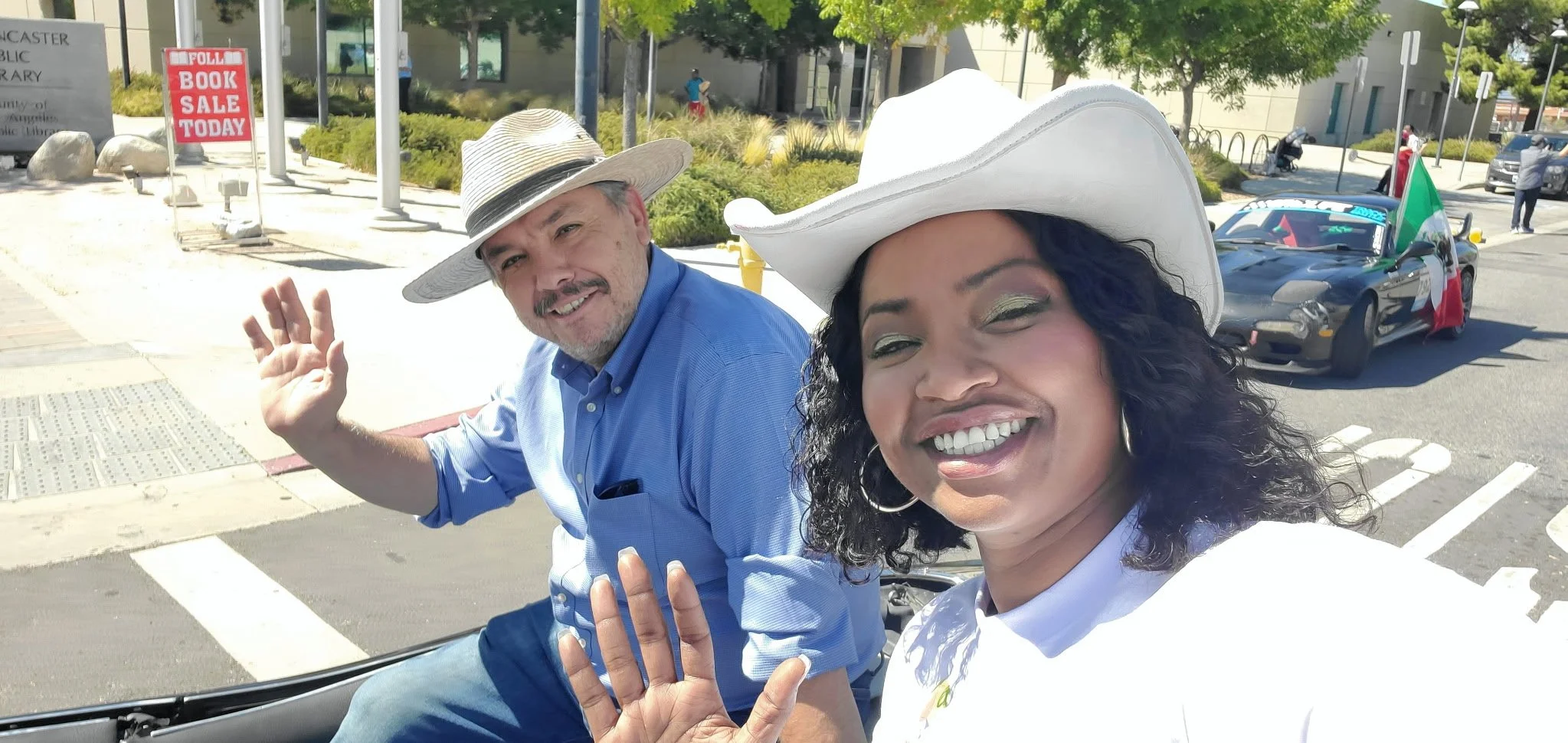 Two people riding in a convertible car, smiling and waving at the camera. The woman is wearing a white hat and has curly black hair, the man is wearing a tan fedora and a blue shirt. Behind them, there is a police car with an Italian flag and people on the sidewalk.