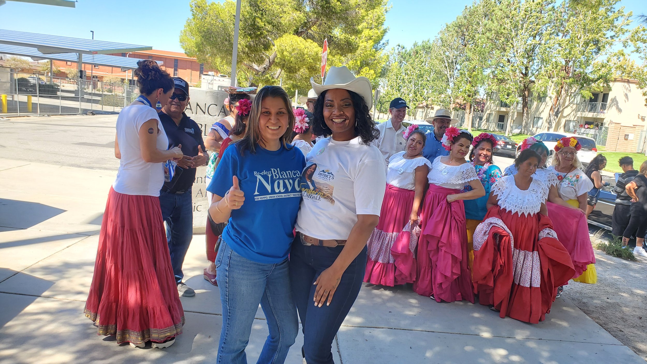 People dressed in traditional Mexican attire at a cultural event, with some women wearing colorful skirts and floral headbands, and two women in the foreground smiling and posing for the photo.
