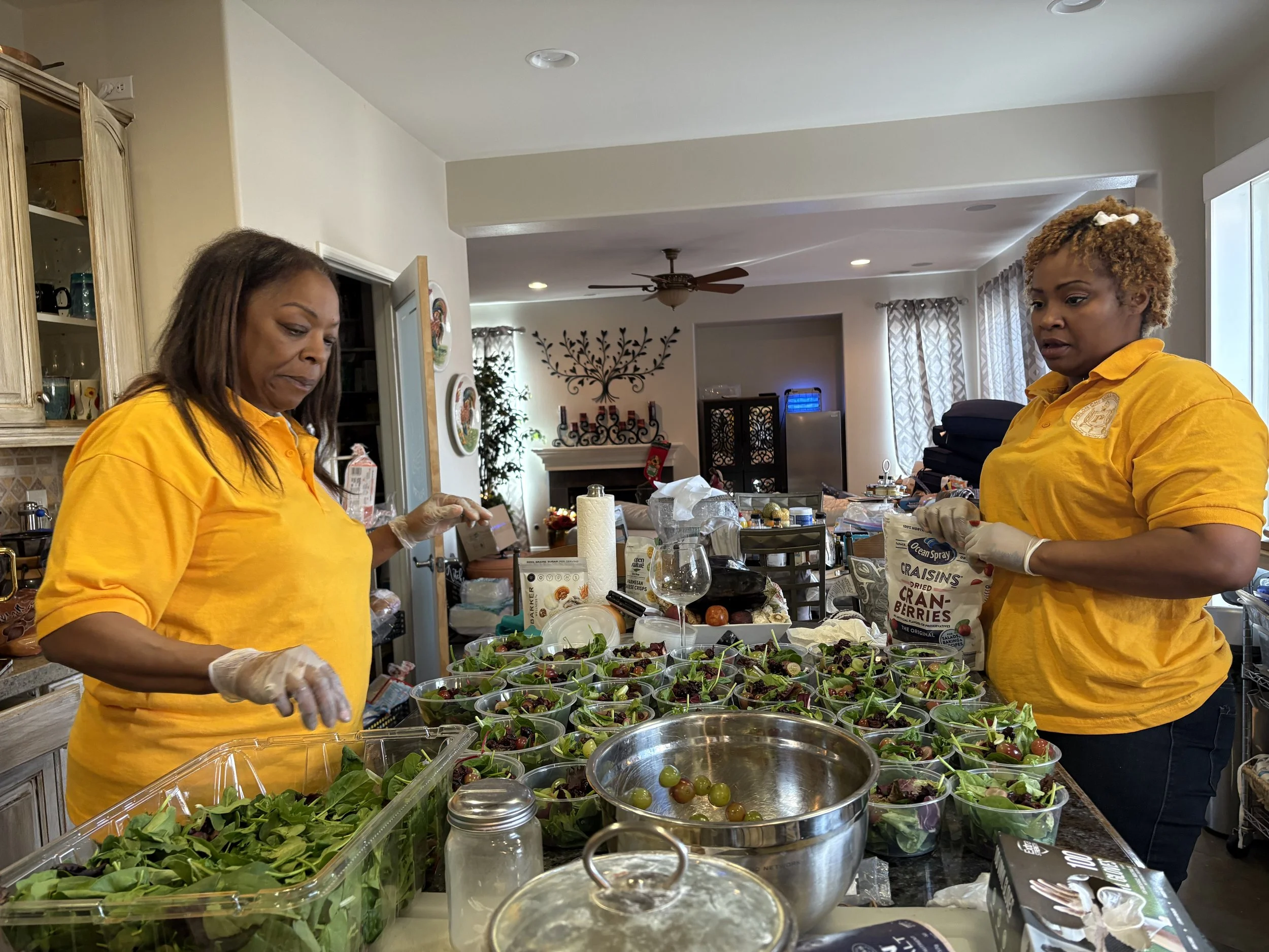 Two women in yellow shirts preparing salads in a home kitchen, surrounded by bowls of mixed greens, cherry tomatoes, and other salad ingredients.