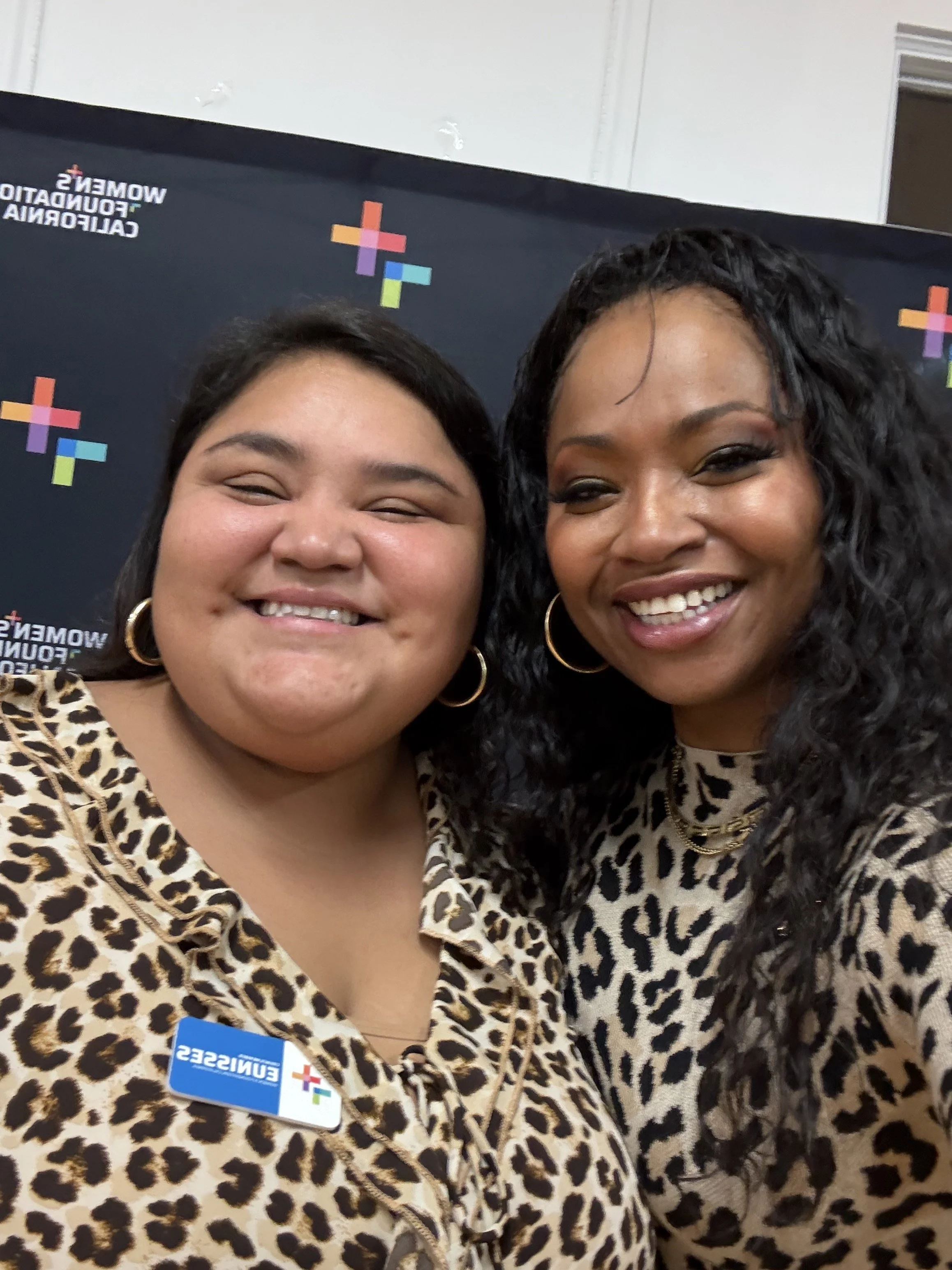 Two women smiling at a conference, wearing leopard print outfits and conference badges with a background that includes colorful crosses and the words 'WOMEN'S FOUNDATION CALIFORNIA'.