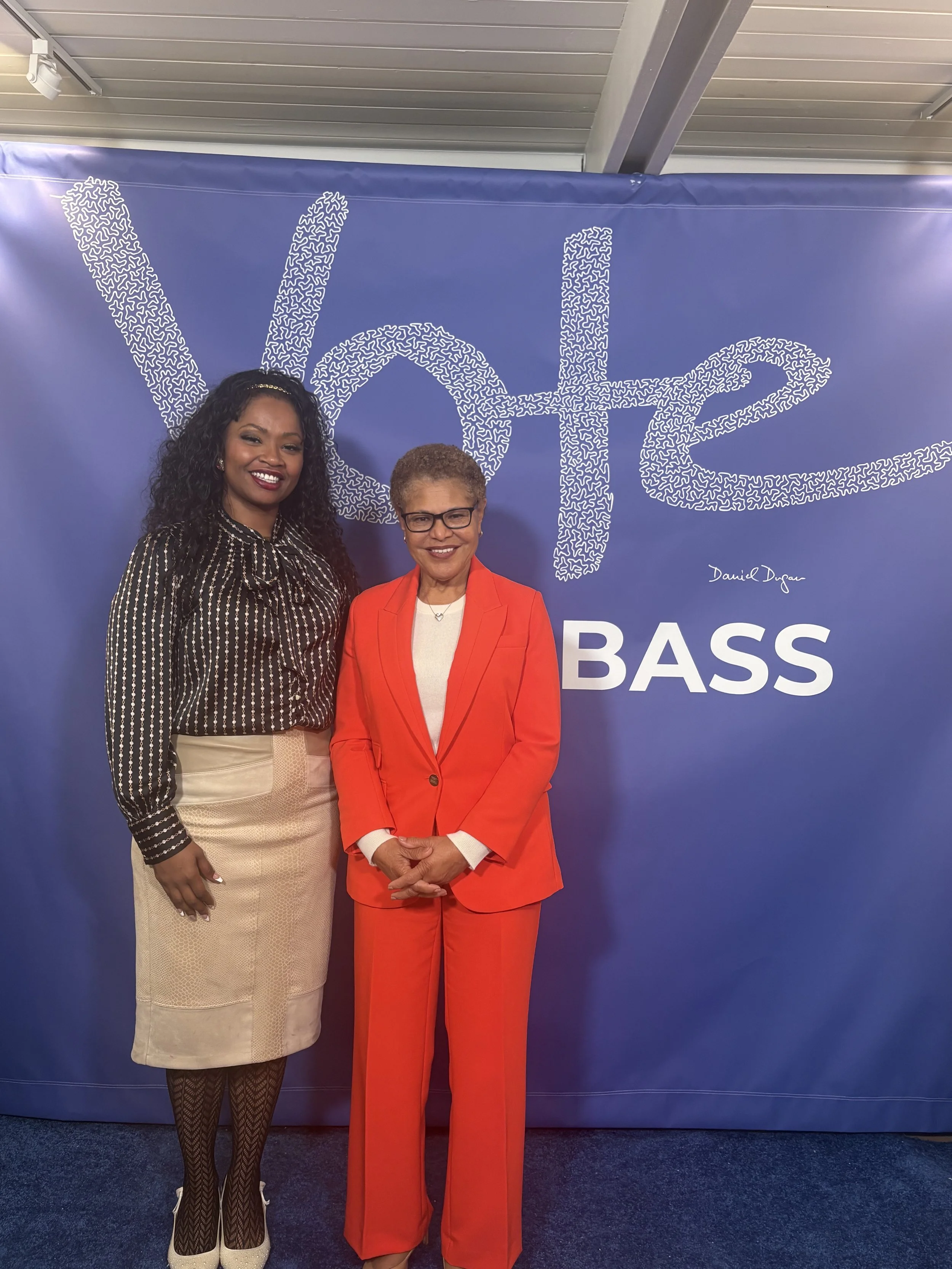Two women standing in front of a blue backdrop with the word "Val" and part of the word "BASS" visible. The woman on the left has dark curly hair, is wearing a striped blouse, beige skirt, and patterned tights, and is smiling at the camera. The woman on the right has short hair, glasses, and is dressed in a bright red suit with a white shirt, also smiling at the camera.