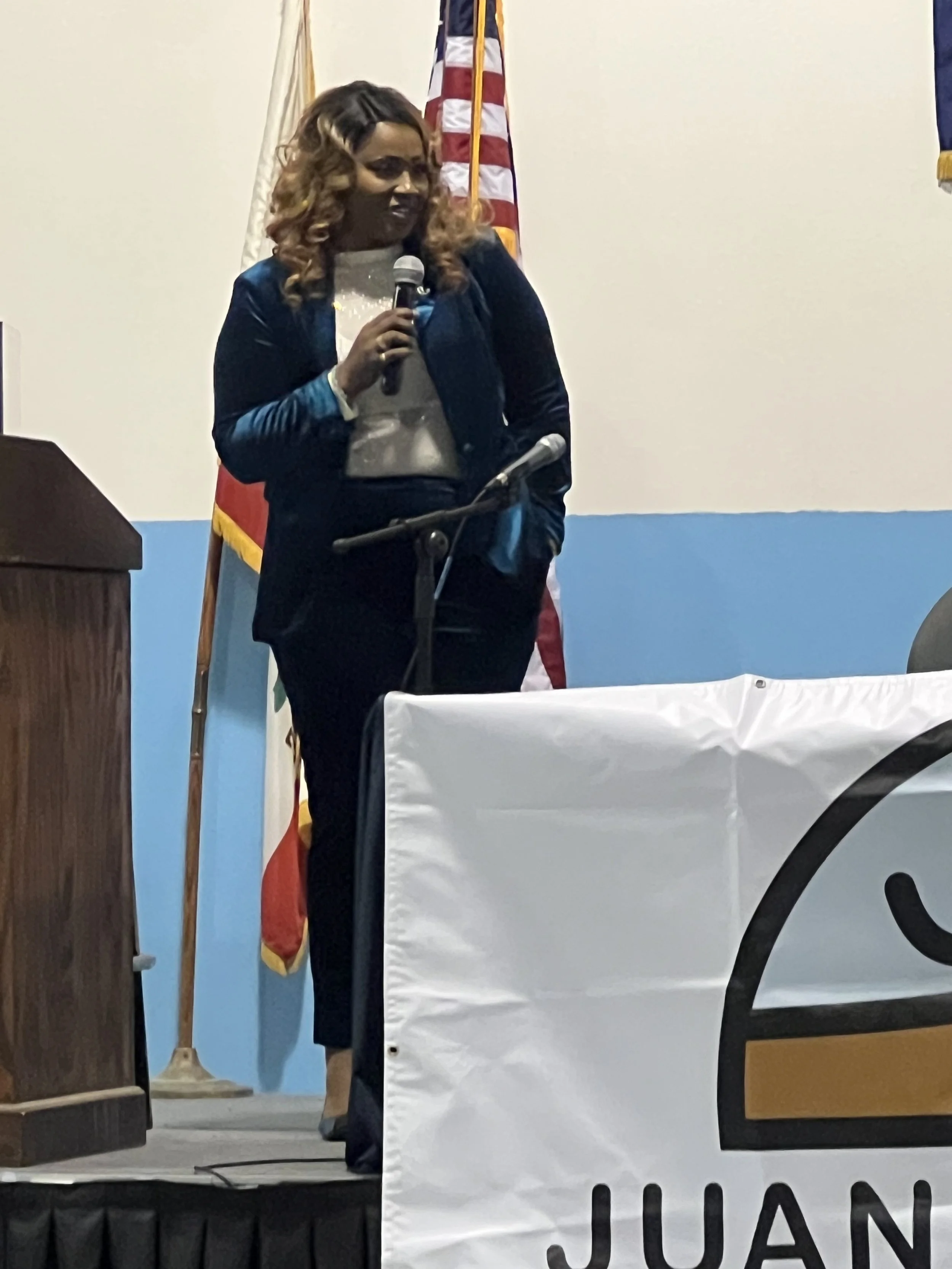 A woman with curly hair holding a microphone speaking at an event, standing in front of American and other flags.