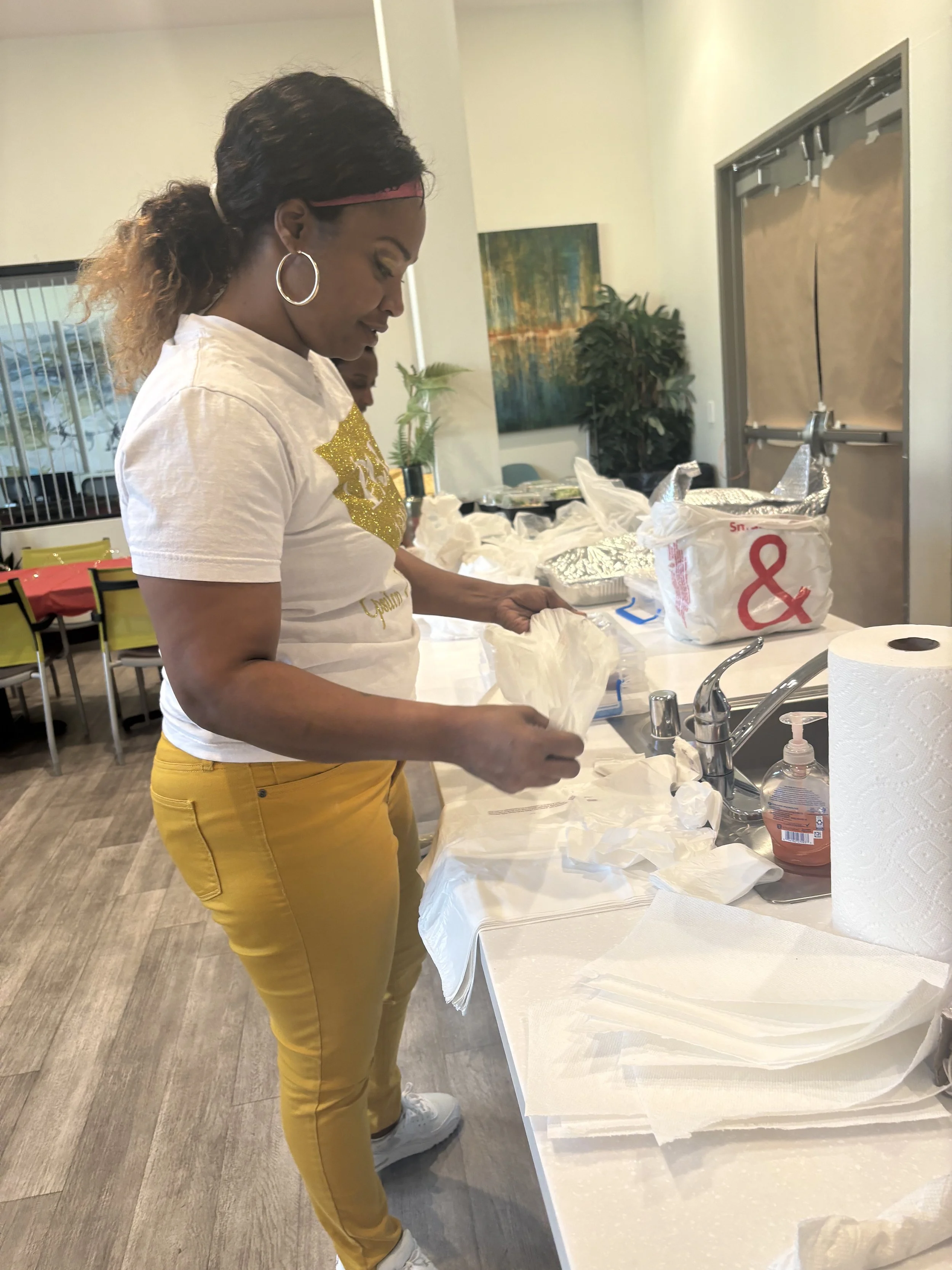 A woman with hoop earrings, wearing a white T-shirt with gold lettering, yellow pants, and white shoes, is standing at a kitchen counter. She appears to be preparing food or handling paper towels, with aluminum foil and a plastic bag on the counter. In the background, there are chairs, a painting, and a large potted plant.