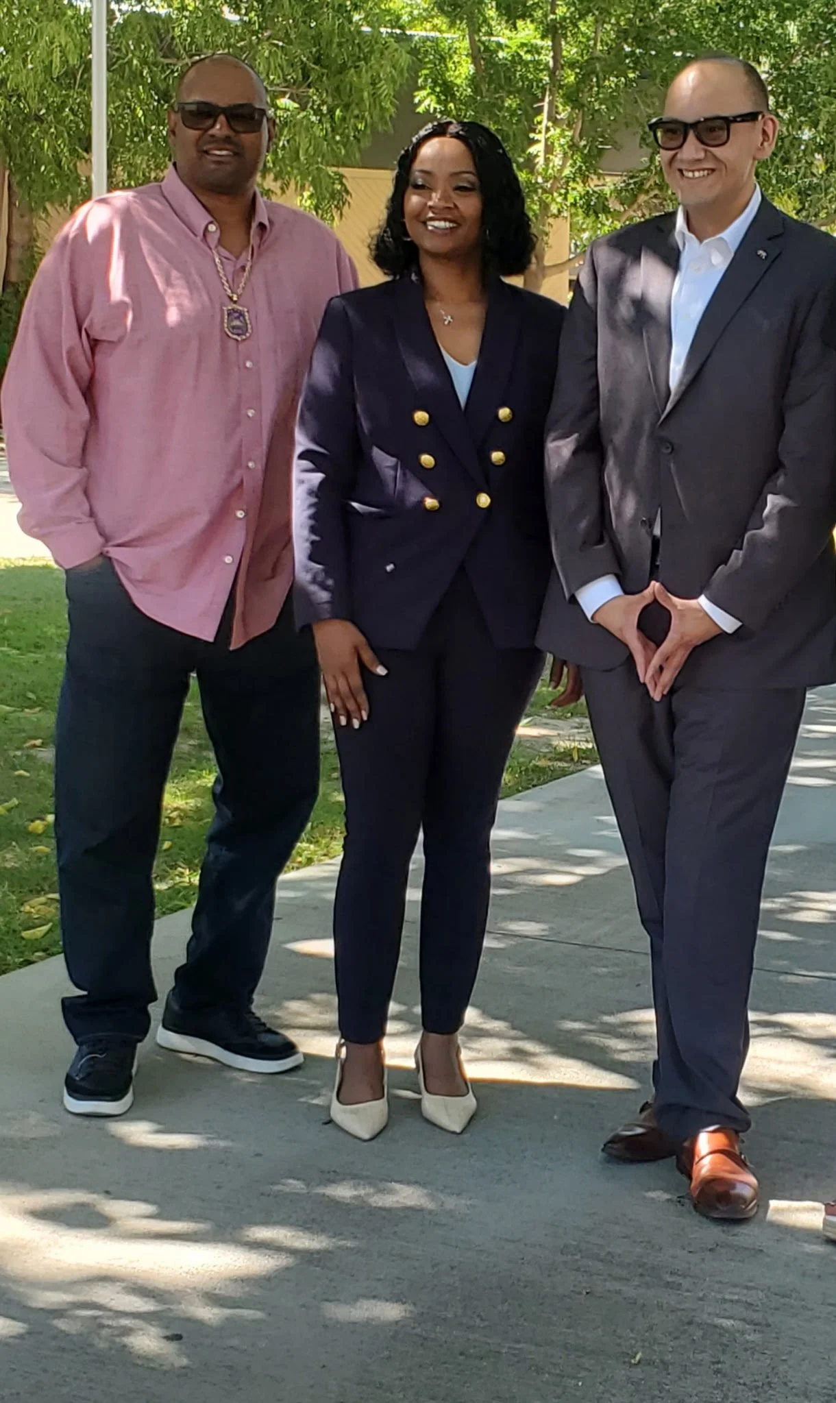 Three people standing outdoors on a sidewalk, posing for a photo, with trees and green foliage in the background.